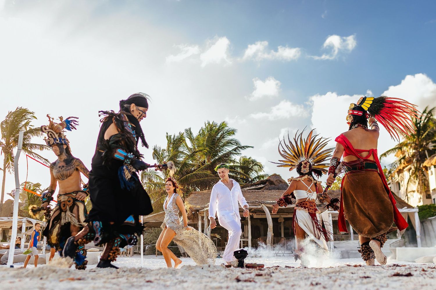 Sarah and Evish laugh while ceremonial participants move around them on the sand.