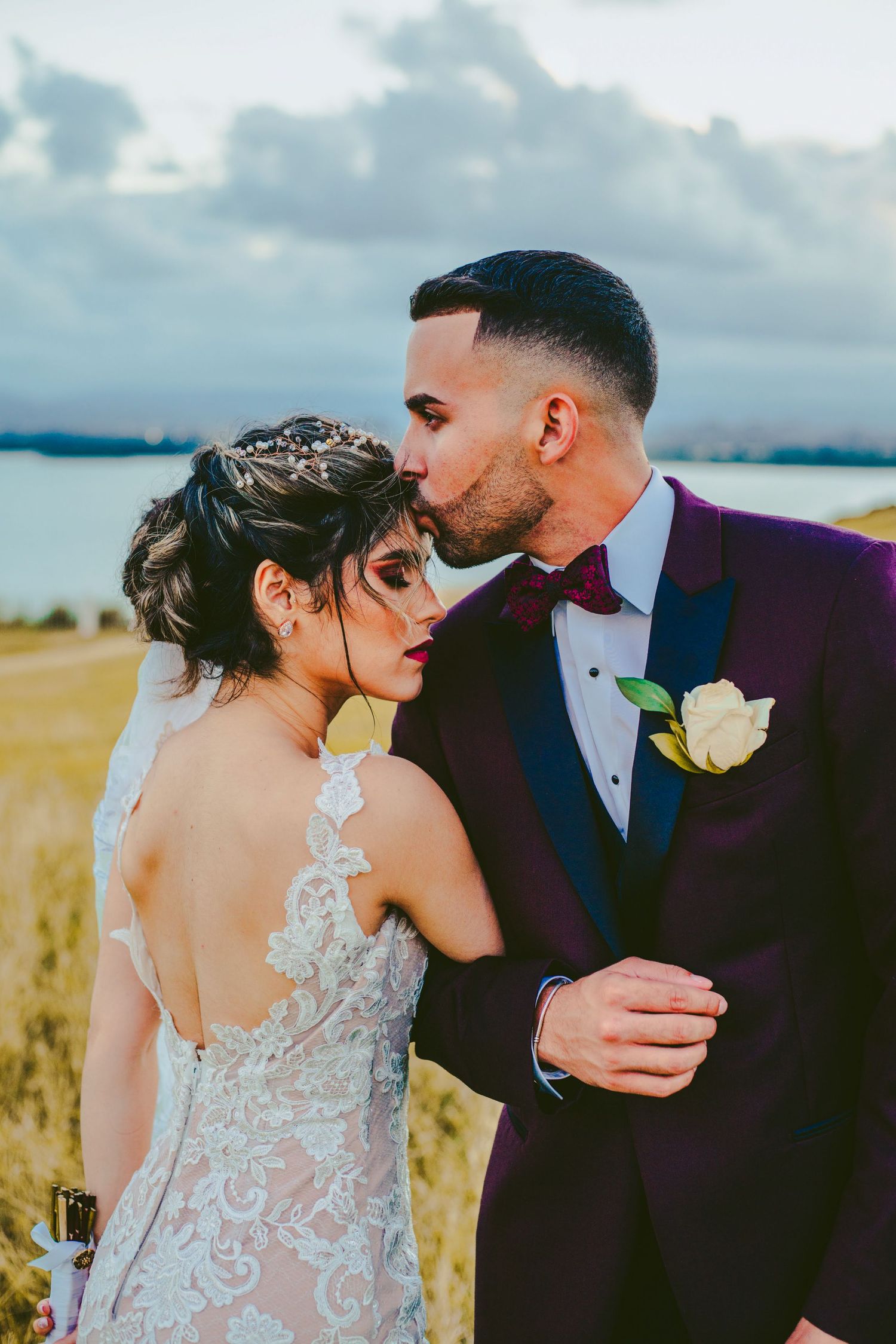 Groom in purple suit kissing bride on cheek in an outdoor field setting.
