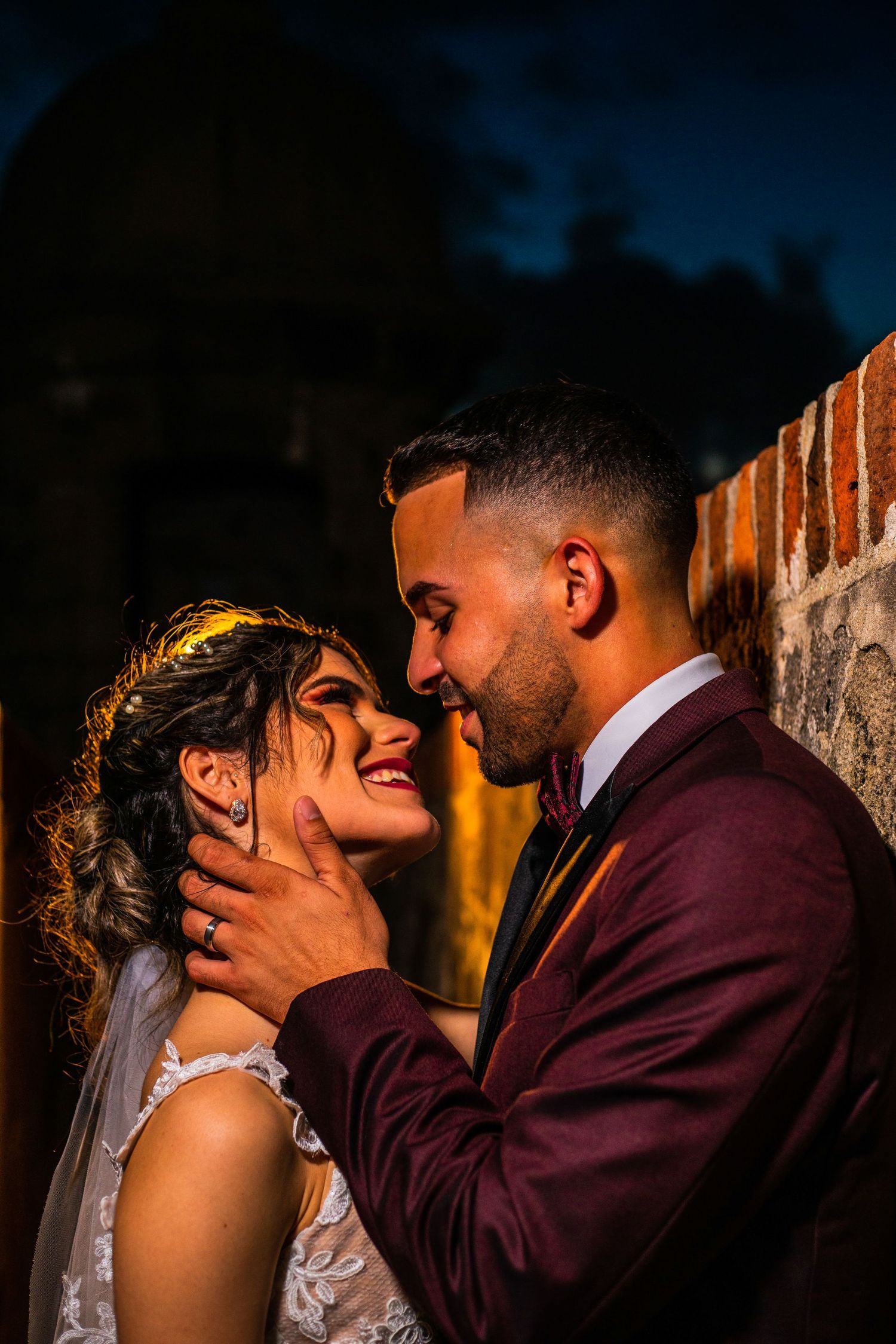 Groom in burgundy suit tenderly holding bride's face near brick wall at night.