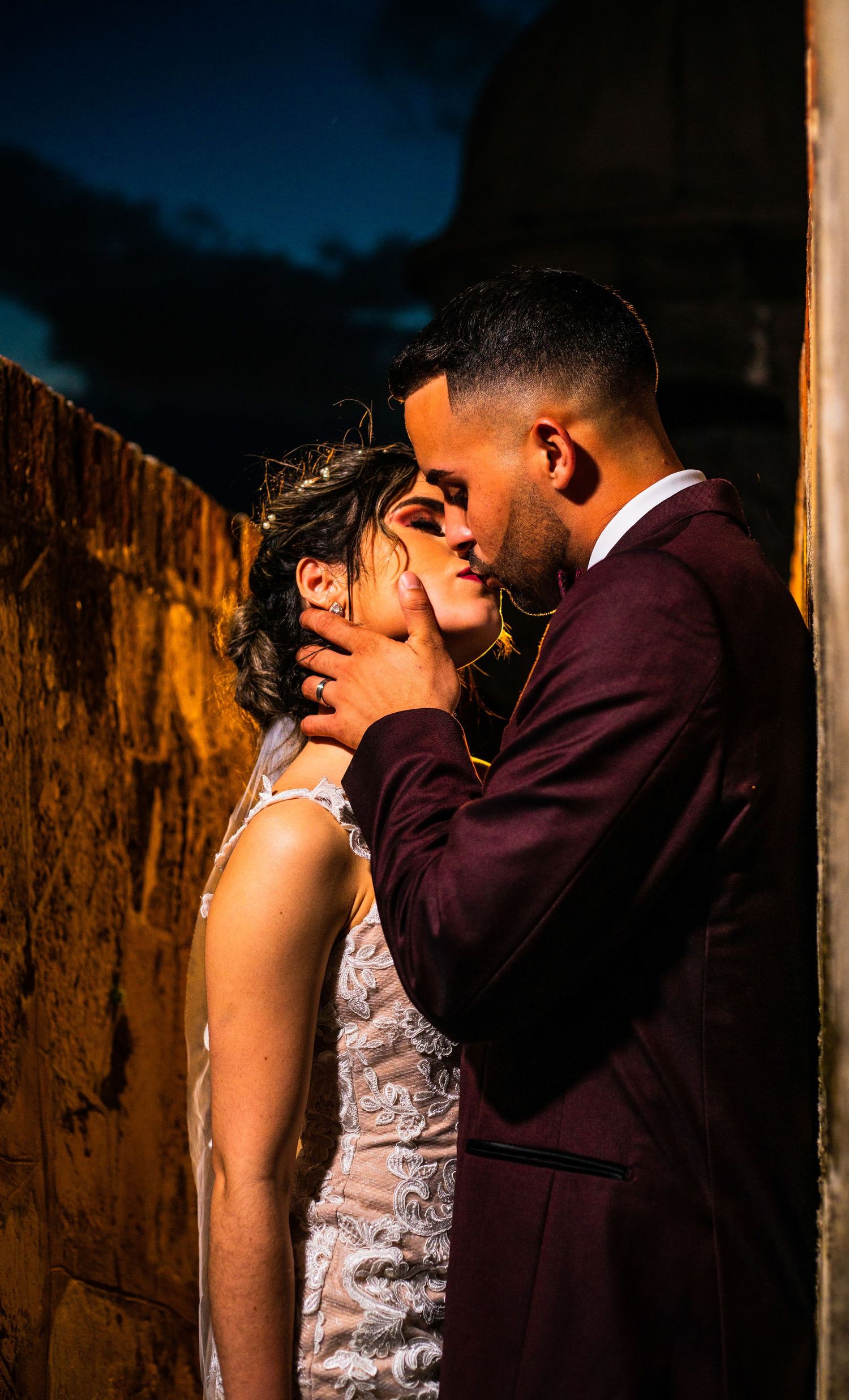 Bride and groom sharing a passionate kiss against a rustic wall at night.