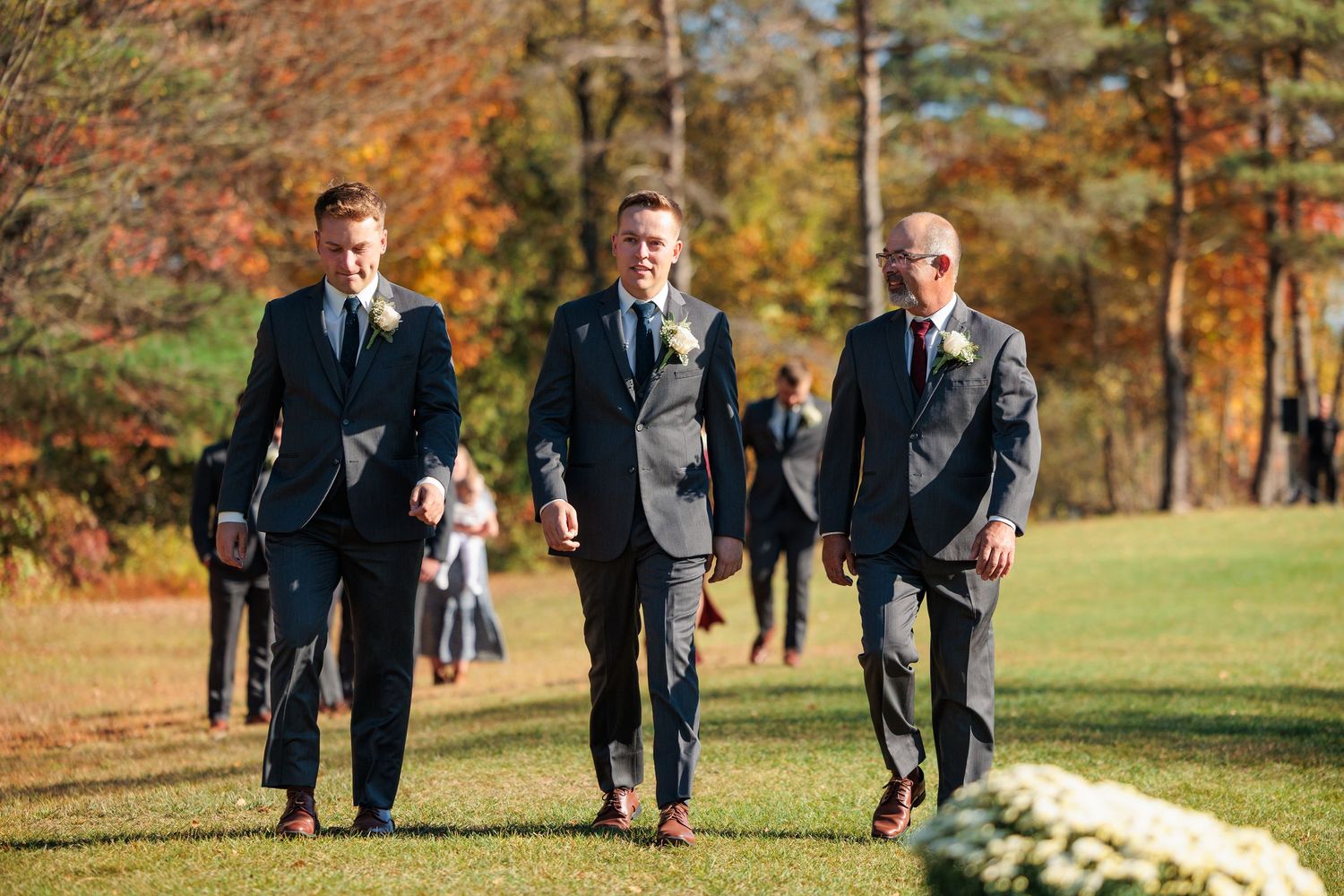 Groomsmen in dark suits walking across a grassy field during autumn.
