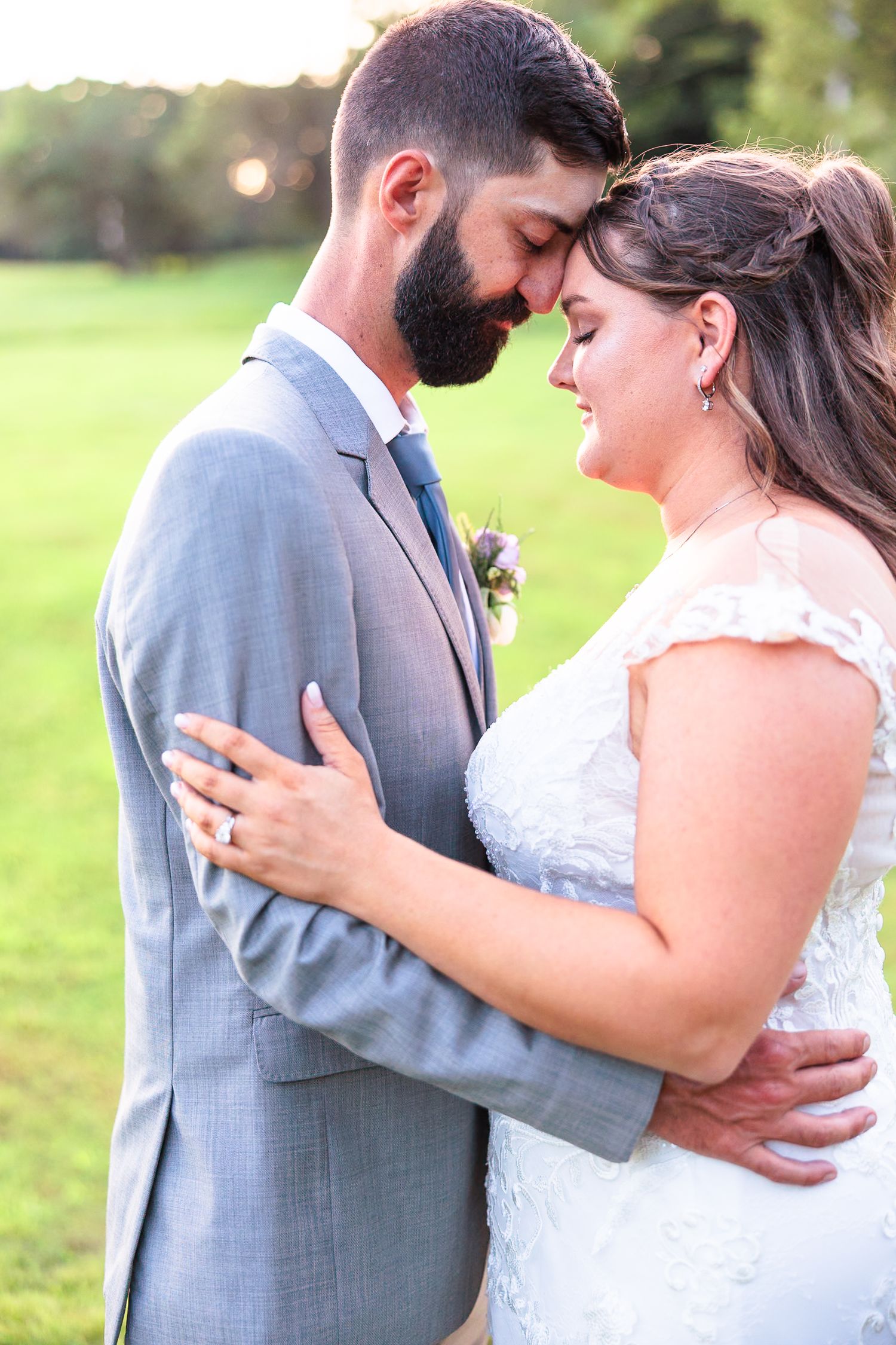 Bride and bearded groom in gray suit touching foreheads tenderly in golden sunset light.