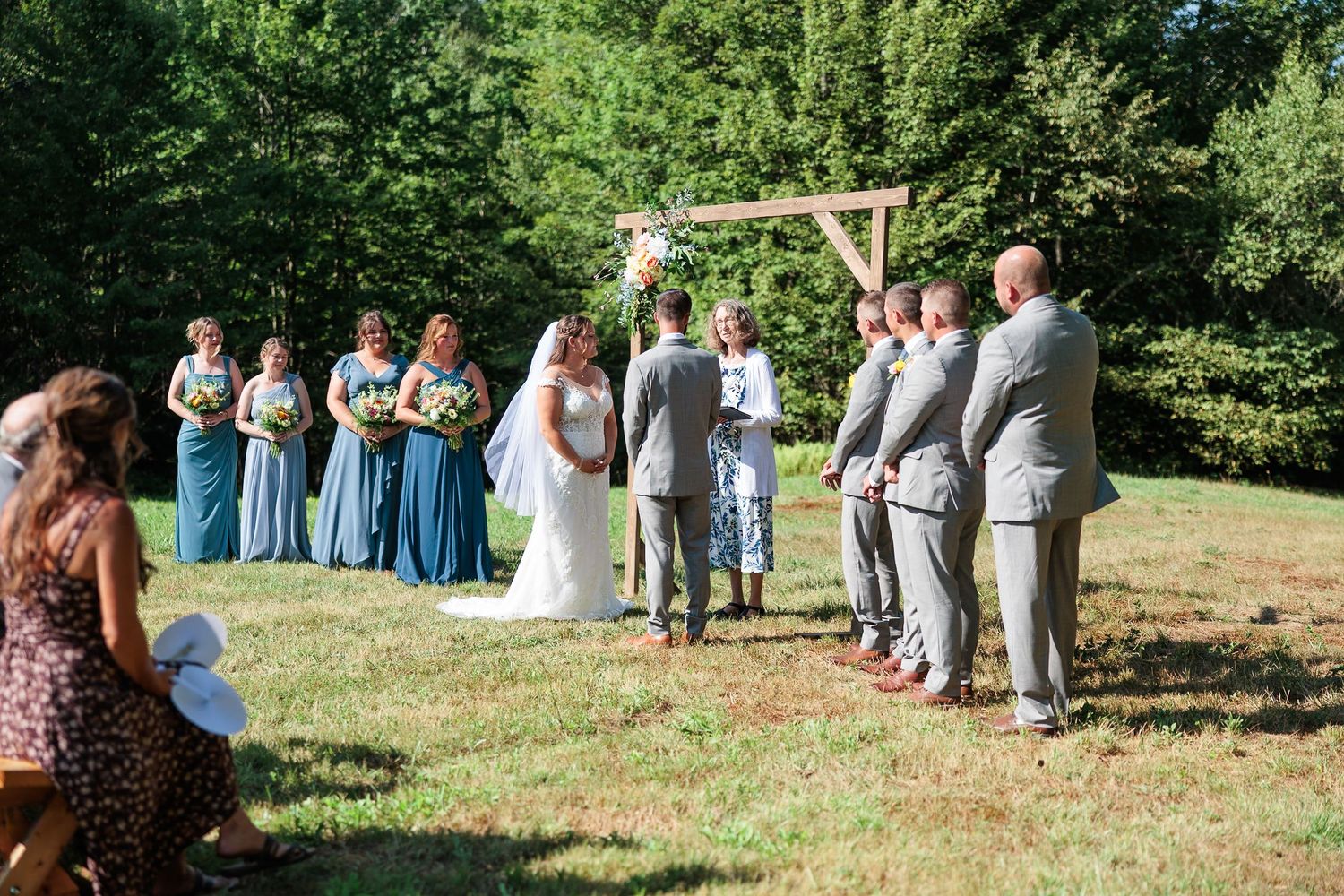 Wedding party stands at rustic wooden arch altar during sunny outdoor ceremony with bridesmaids and groomsmen.