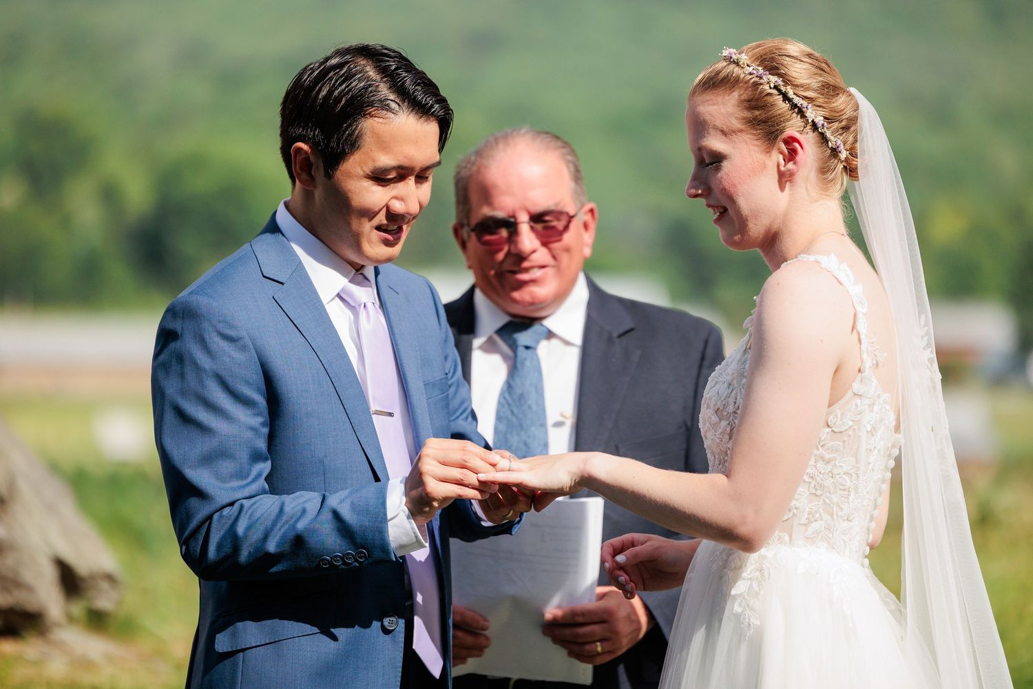Groom places wedding ring on bride's finger during outdoor ceremony with officiant.