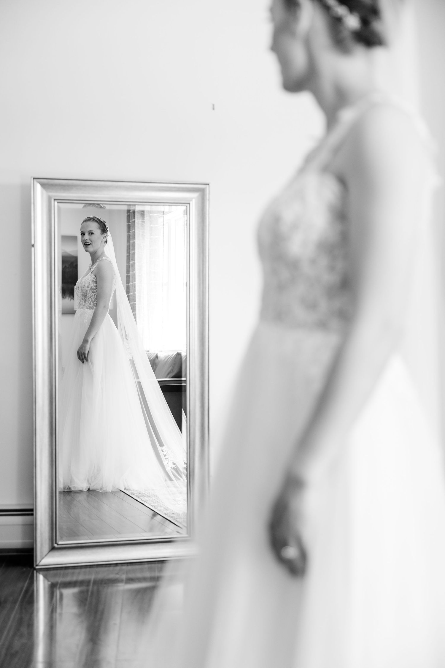 Black and white portrait of a bride in a lace wedding gown reflected in a full-length silver mirror.