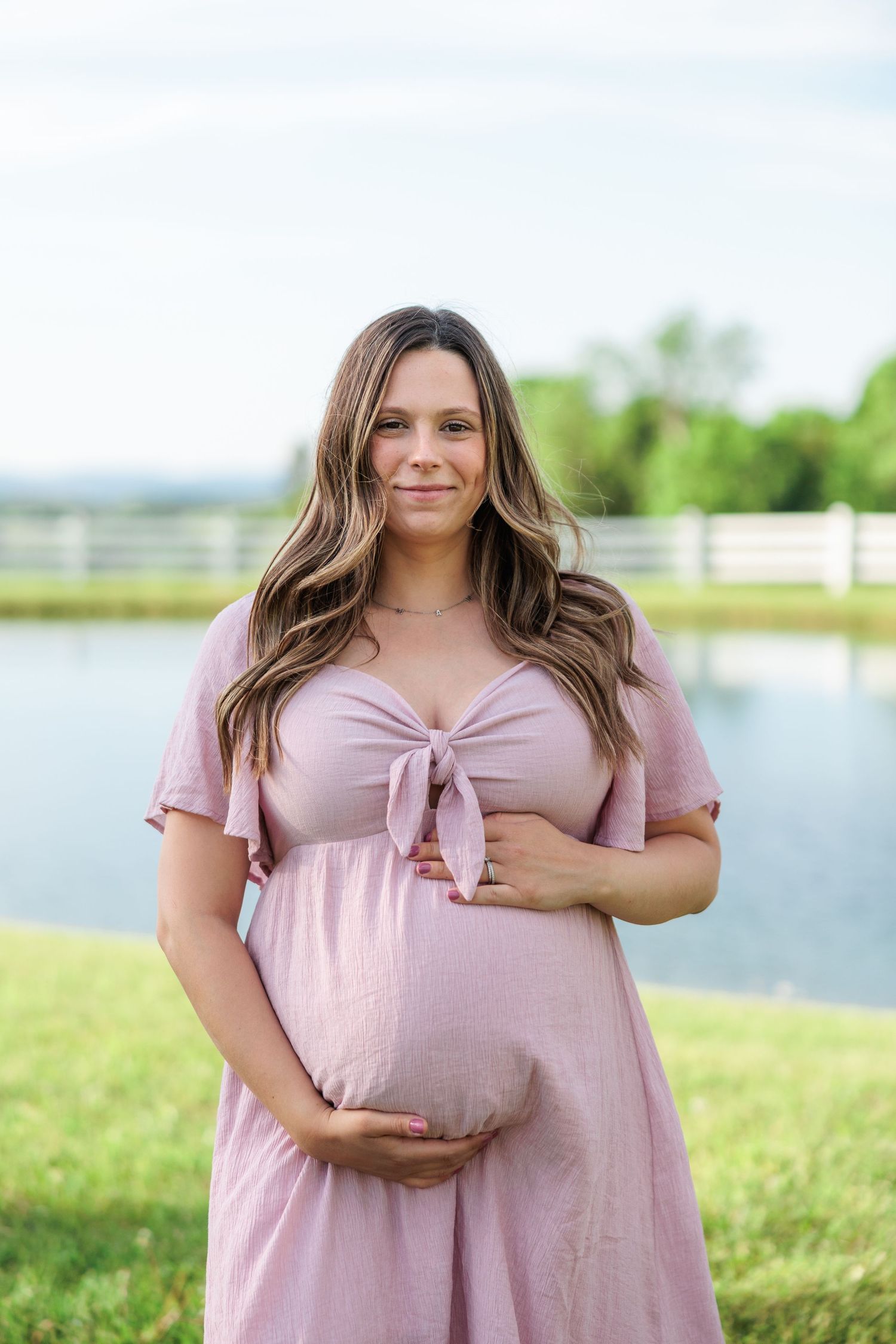 Smiling pregnant woman in pink dress holding baby bump near a pond.