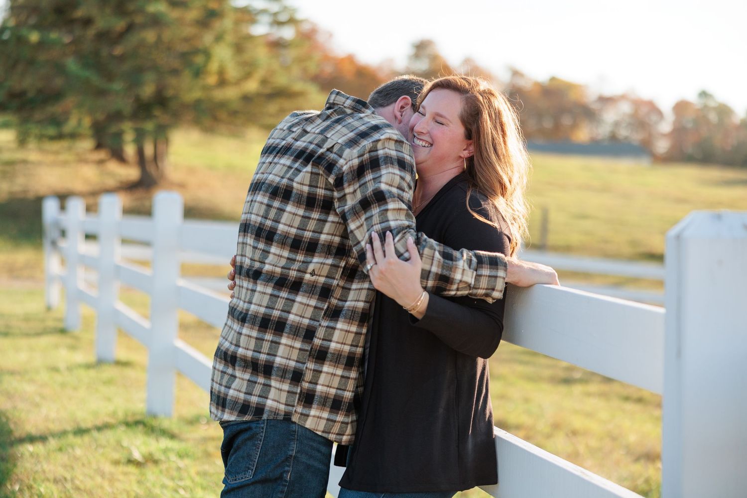 Joyful woman laughing as her partner nuzzles her neck against a white farm fence at sunset.