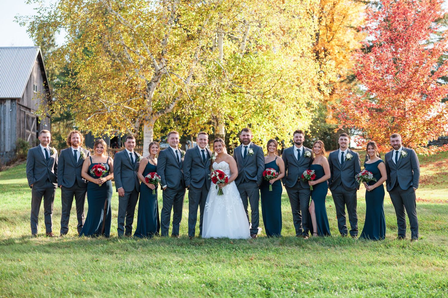 Wedding party in gray suits and teal gowns pose together on green lawn with colorful autumn trees behind.