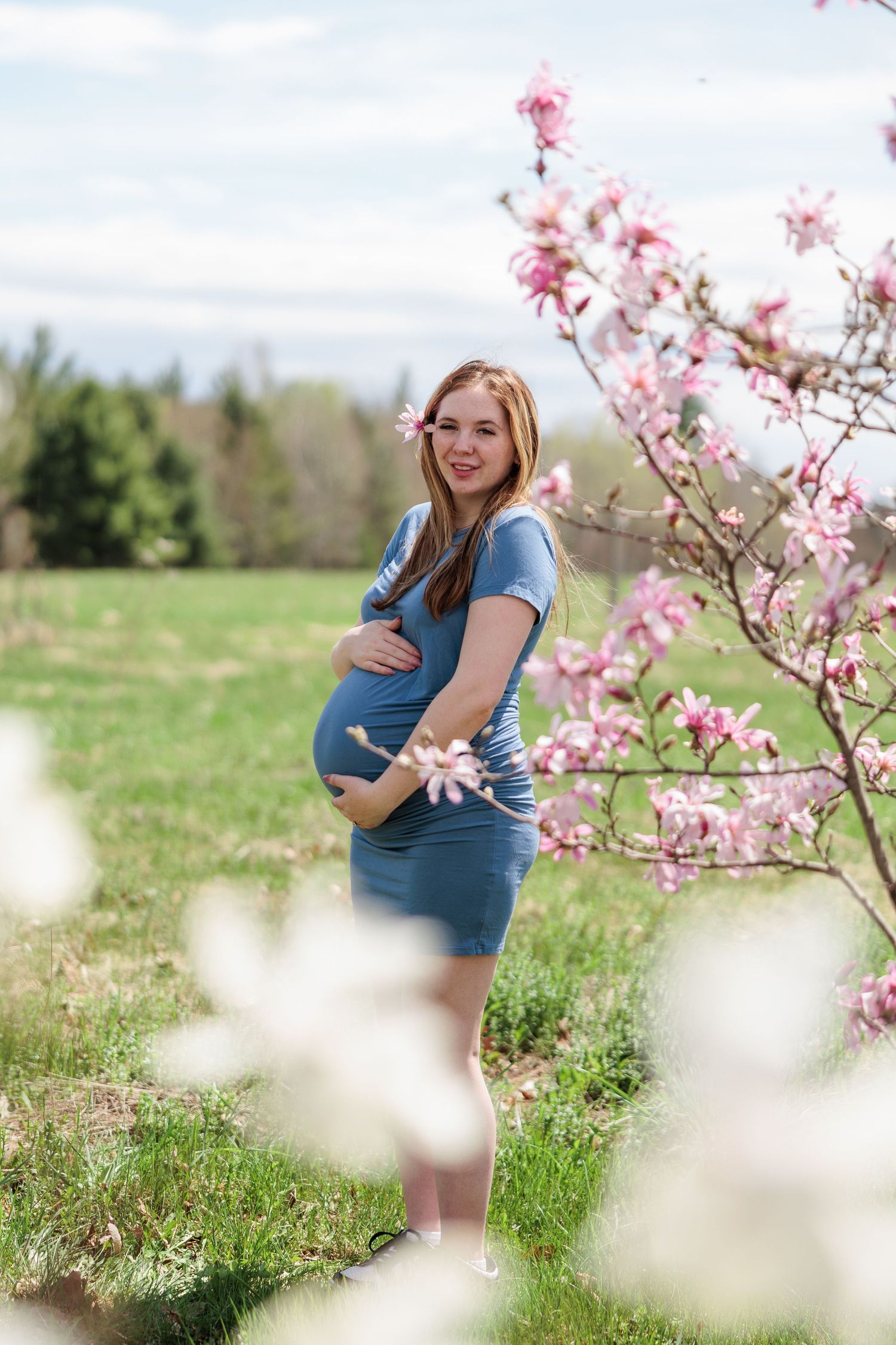 Smiling pregnant woman in blue dress cradling bump beside pink magnolia blossoms in a sunny green field.
