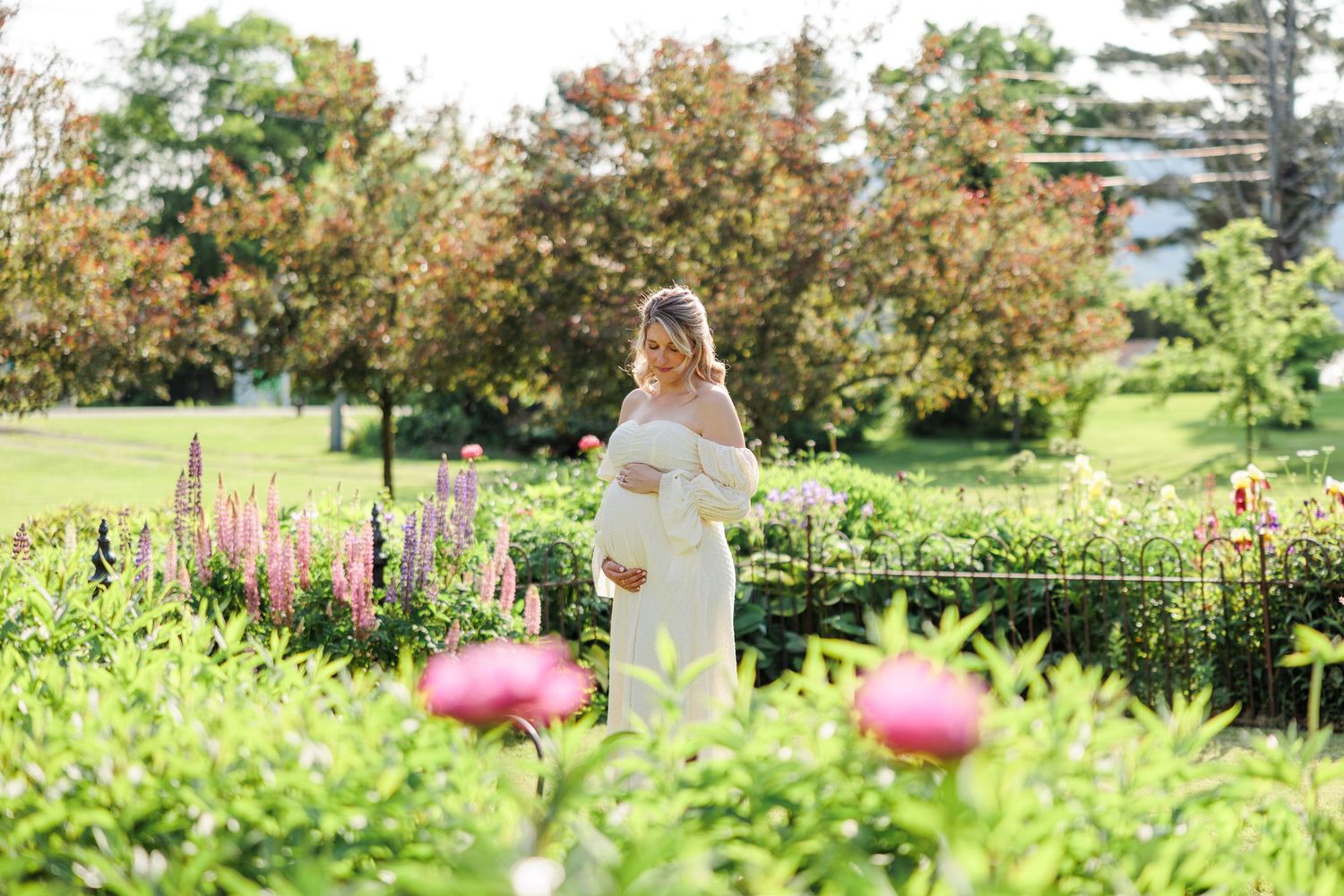 Pregnant woman in white dress standing in lush garden surrounded by colorful blooming flowers.