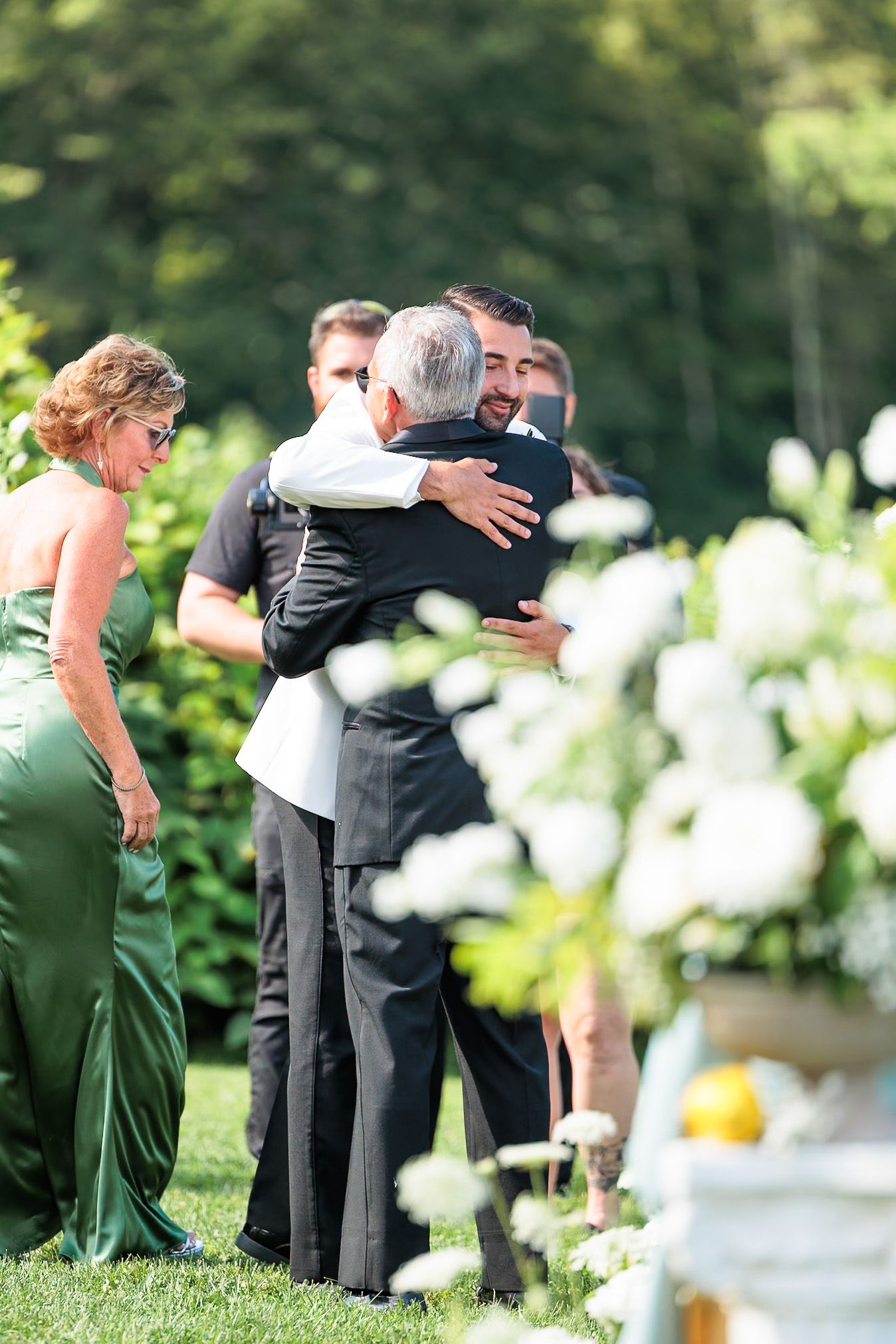 Two men sharing an emotional hug at an outdoor wedding ceremony surrounded by guests.