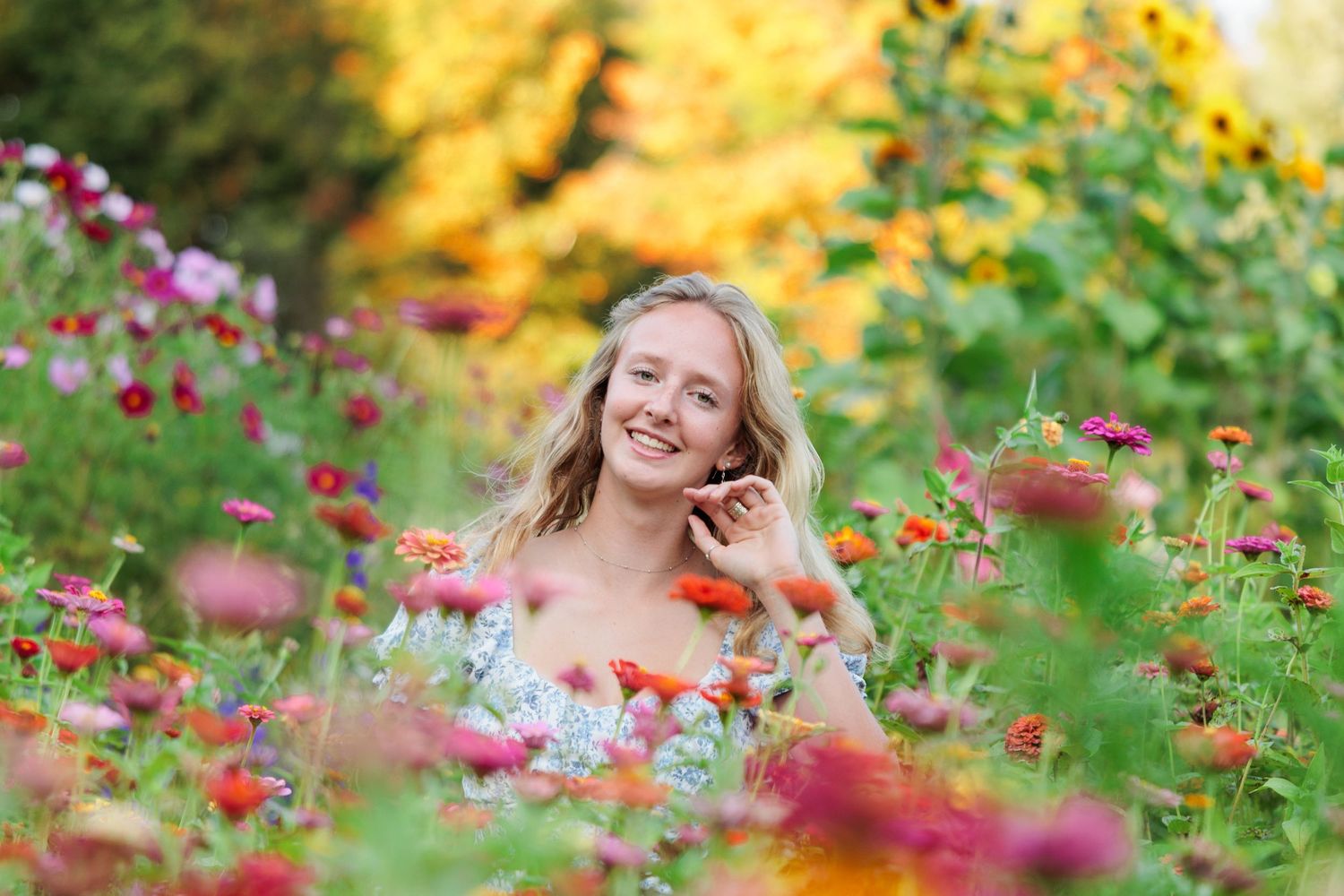 Smiling young woman with blonde hair talking on phone surrounded by colorful zinnia flowers in a garden.