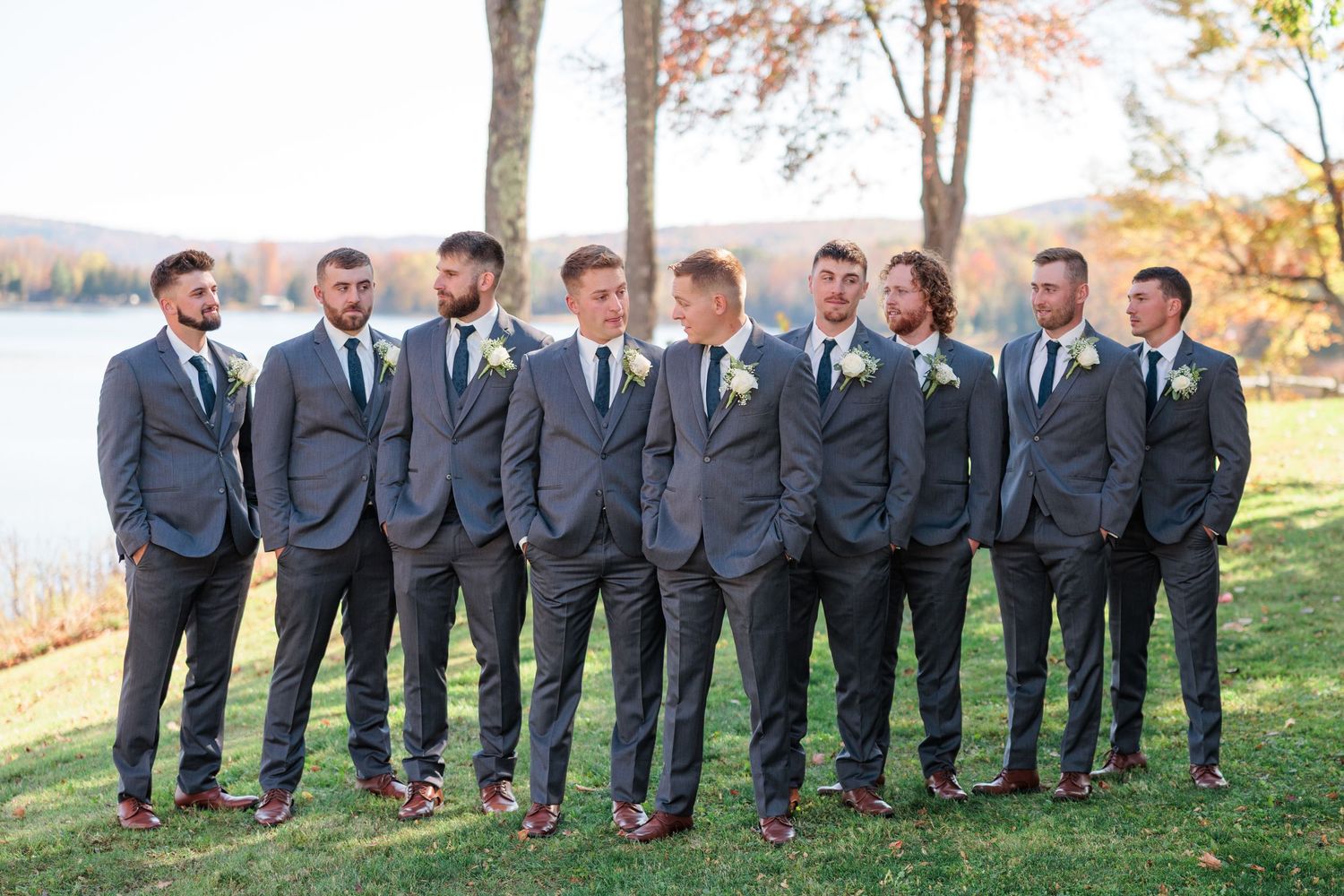 Wedding party in gray suits poses together on a sunny autumn day with fall foliage in the background.