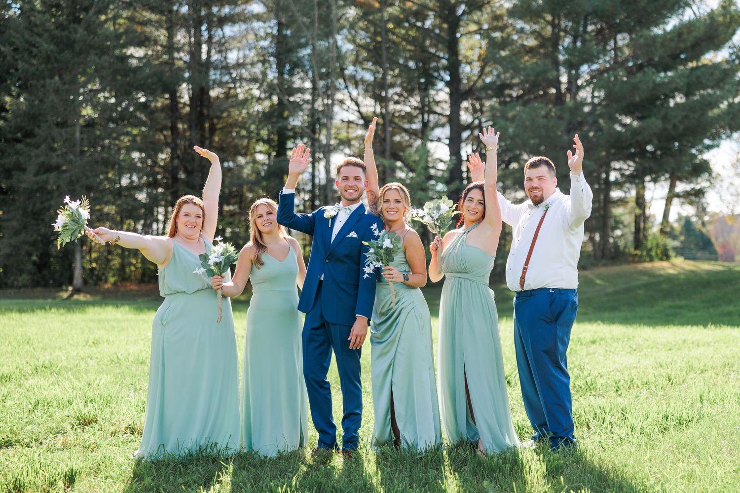 Celebratory wedding party in mint green dresses and navy suits cheering with raised arms on sunny lawn.