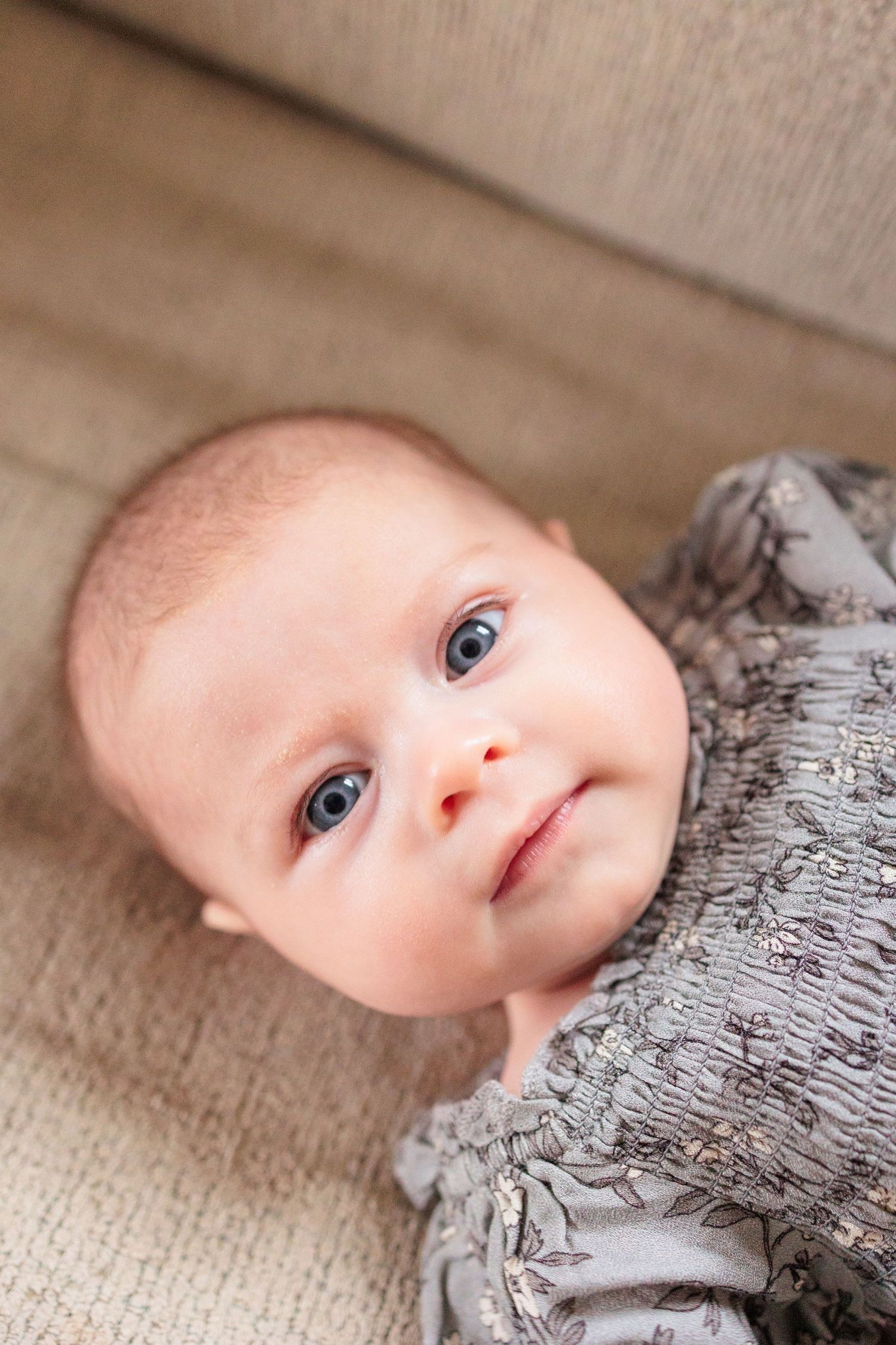 A sweet baby wearing a gray outfit rests on a beige couch cushion.