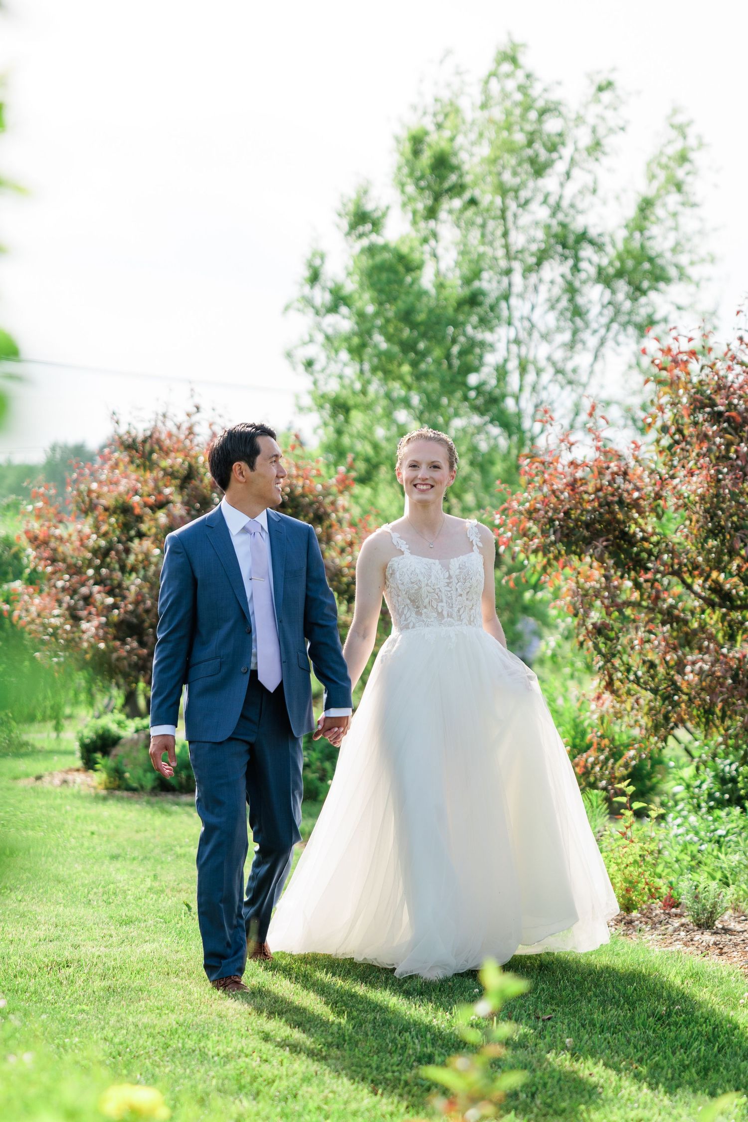 Bride in white tulle gown and groom in navy suit walking hand in hand through a blooming garden.