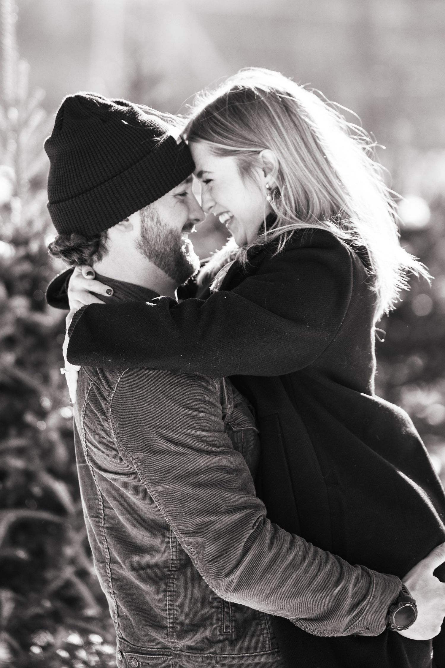 Black and white photo of a couple sharing a joyful embrace, nearly kissing among winter greenery.