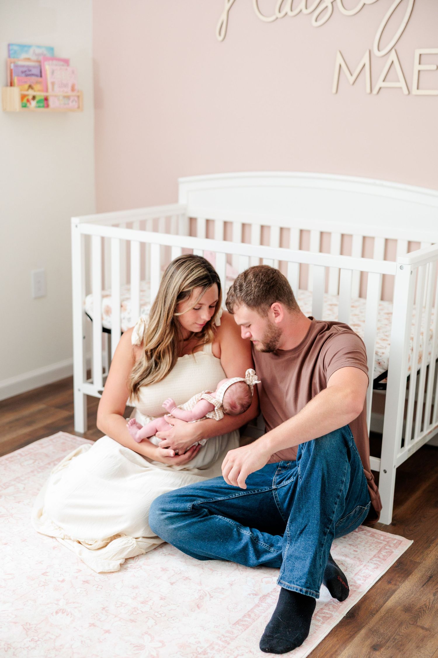 New parents sitting on nursery floor holding newborn baby girl in front of white crib.