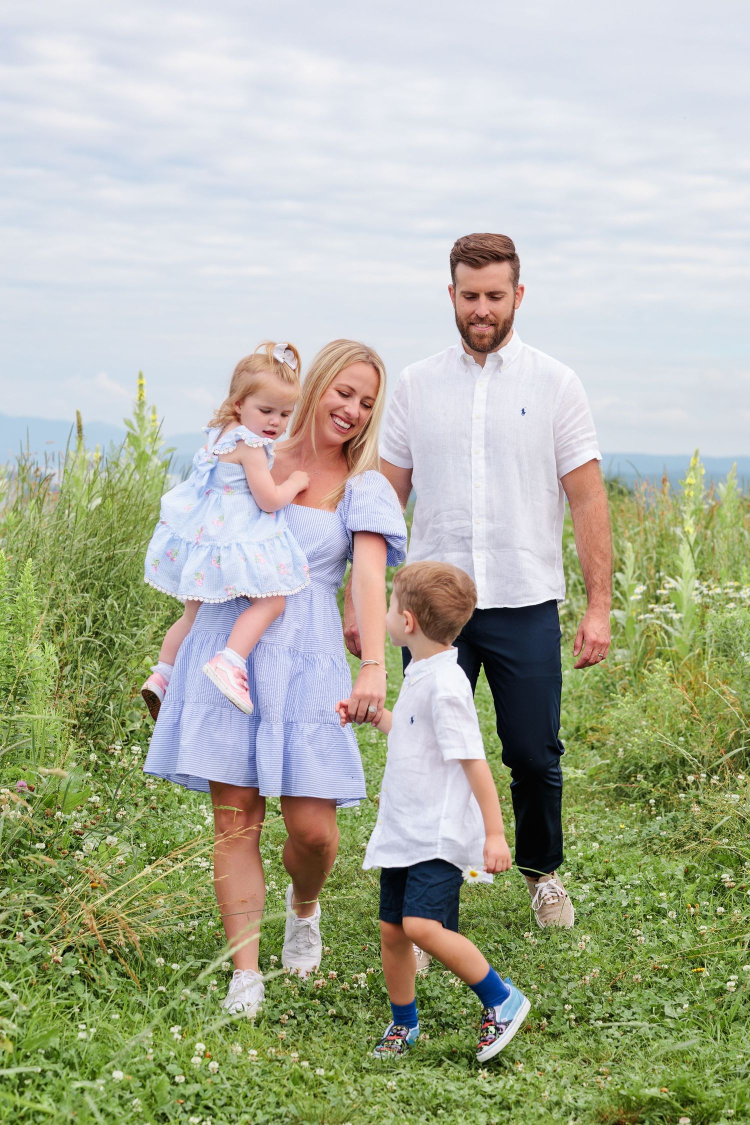 Young family of four walking hand in hand through a lush green wildflower meadow together.