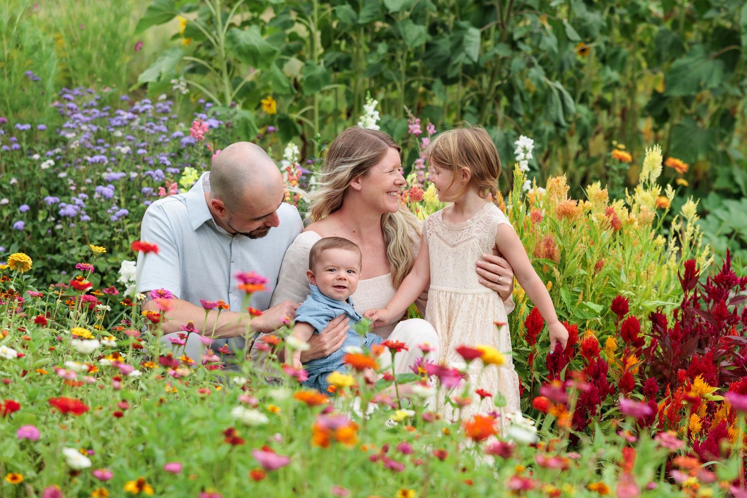 A happy family of four sits together surrounded by vibrant wildflowers in a lush garden.