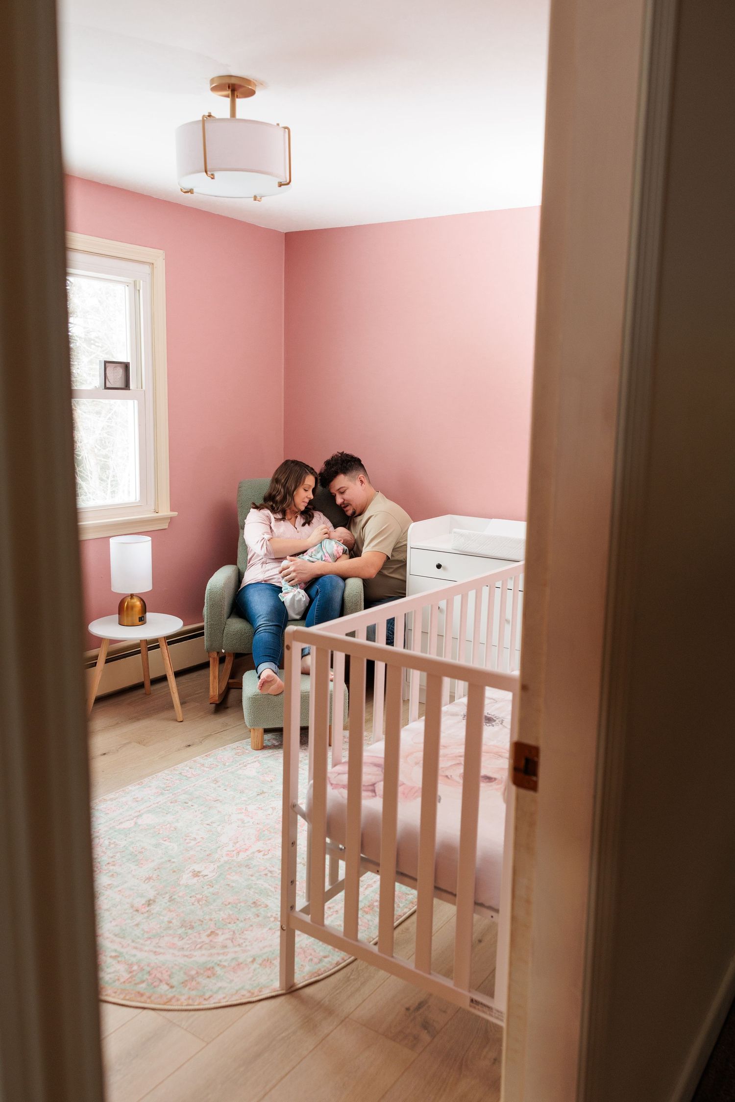 New parents sit together in a pink nursery holding their newborn baby near a white crib.