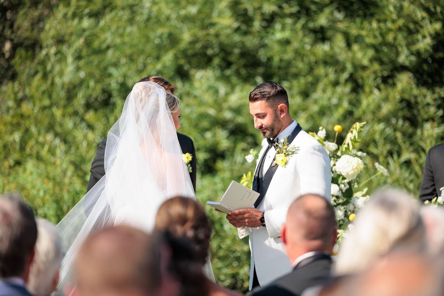 Groom in white tuxedo reading heartfelt wedding vows to veiled bride during outdoor garden ceremony.