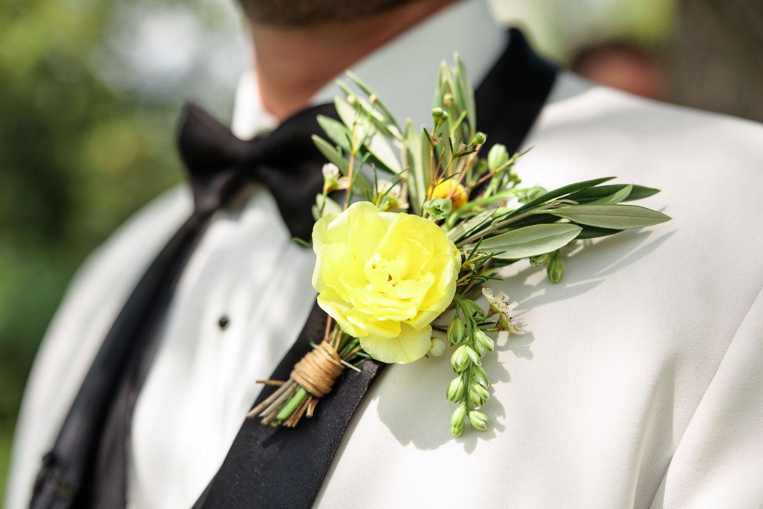 Groom wearing a bright yellow ranunculus boutonniere with greenery on a white tuxedo jacket.