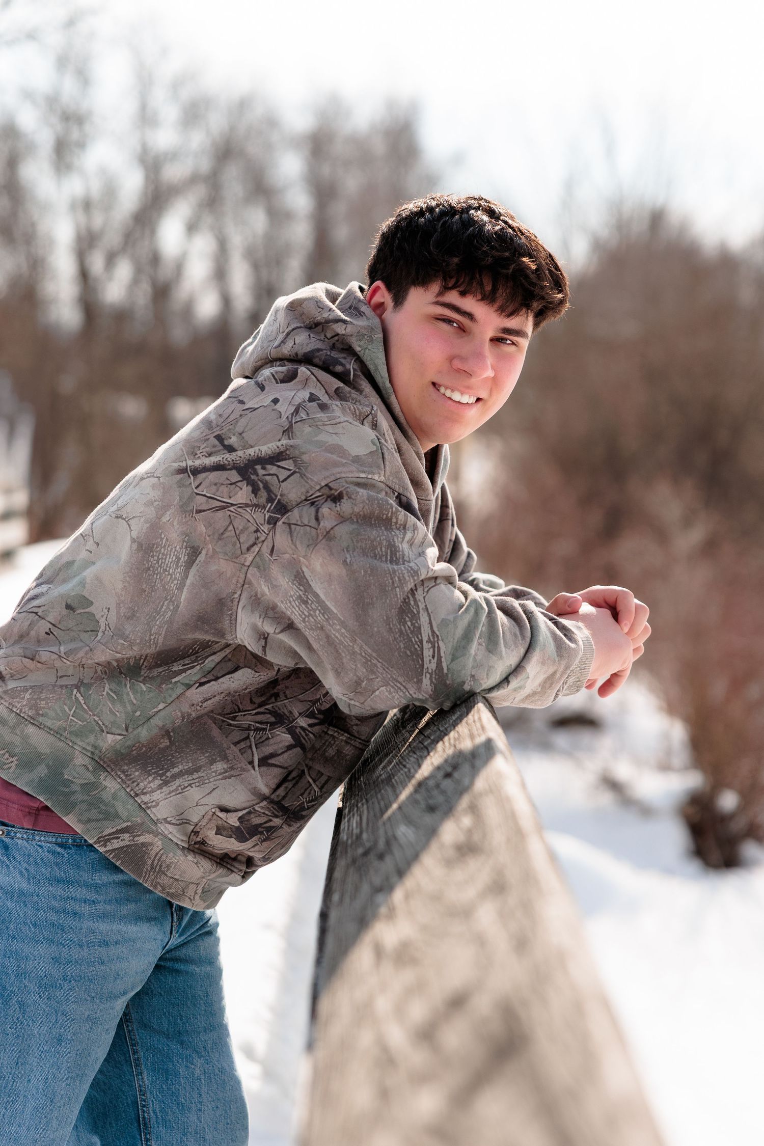 Smiling teenage boy in camo jacket and jeans leaning on a railing outdoors in a snowy winter landscape.