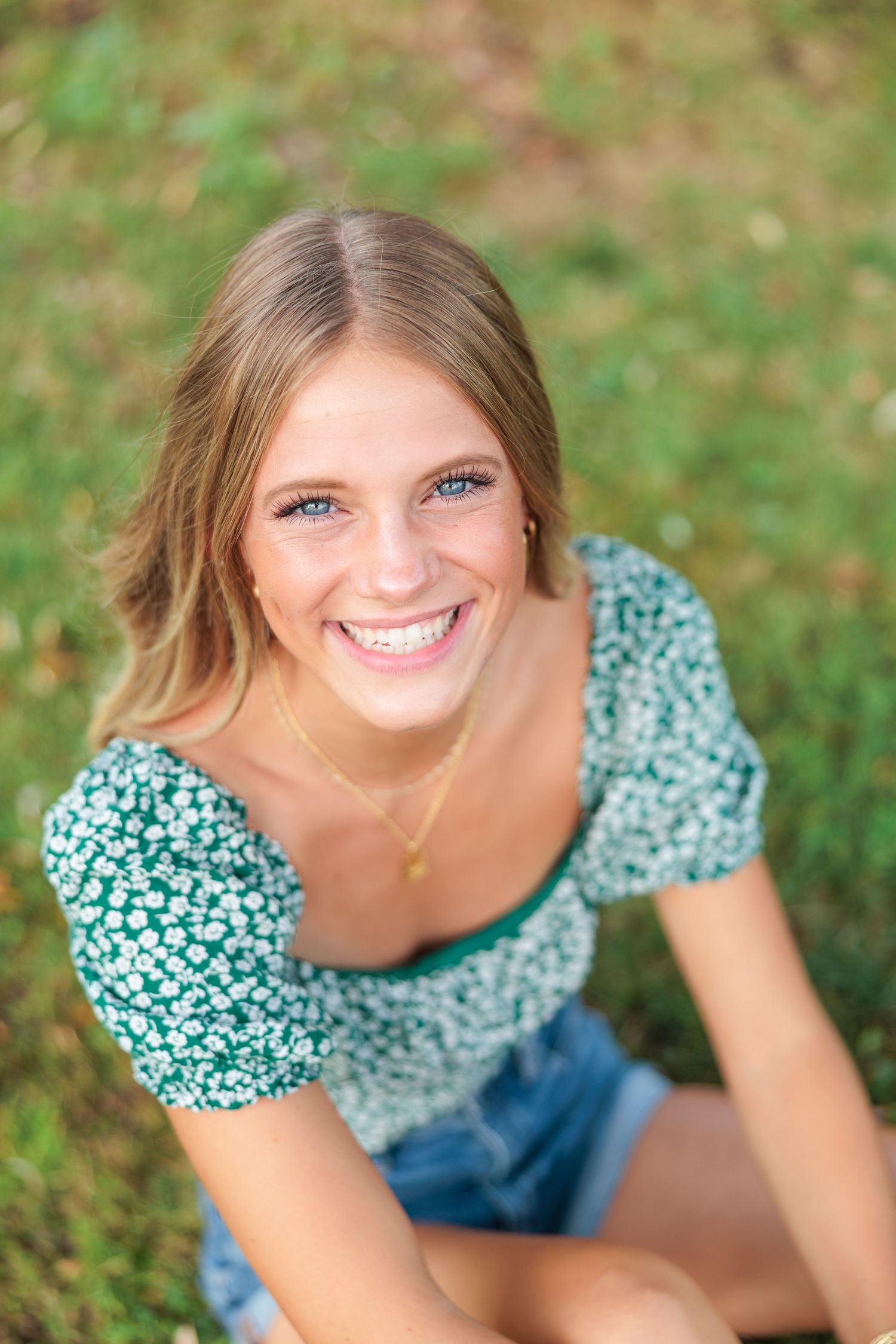 Young woman with blue eyes smiling outdoors, wearing a green floral crop top and denim shorts, sitting on grass.