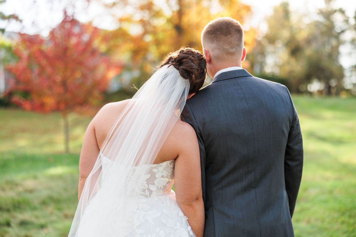 A couple in wedding attire stands close together on a grassy hill during autumn.