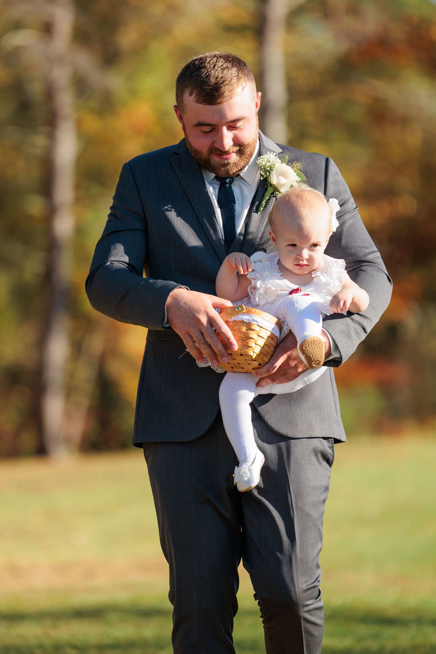 A person in a dark suit holds a small baby wearing white at an outdoor autumn wedding.