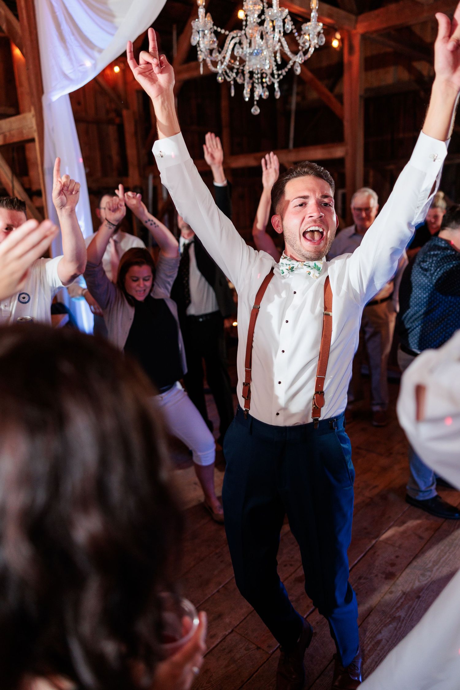 Guest dancing enthusiastically under a crystal chandelier at a barn wedding reception.