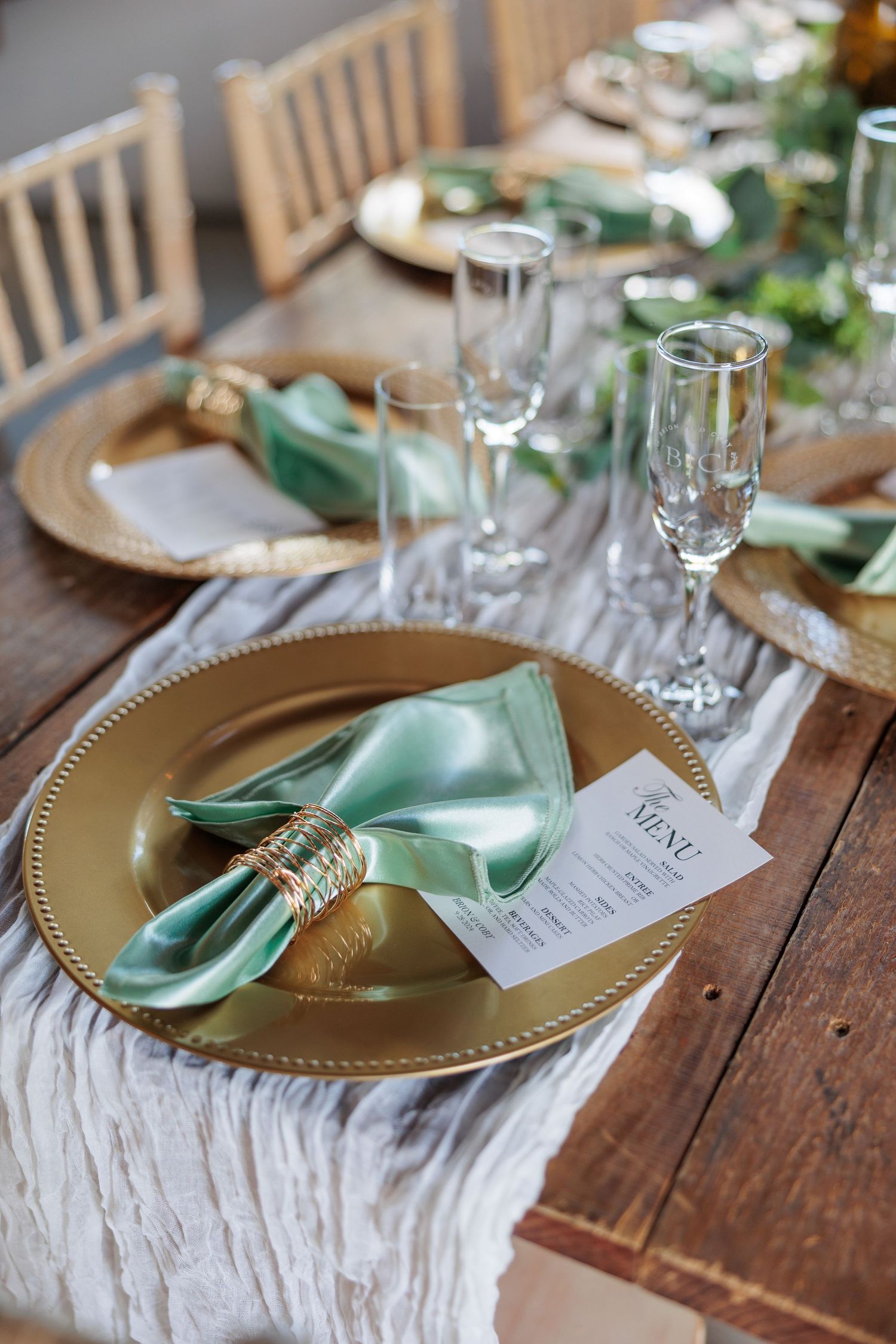Place settings with gold charger plates, mint napkins, and menu cards on rustic wooden tables.