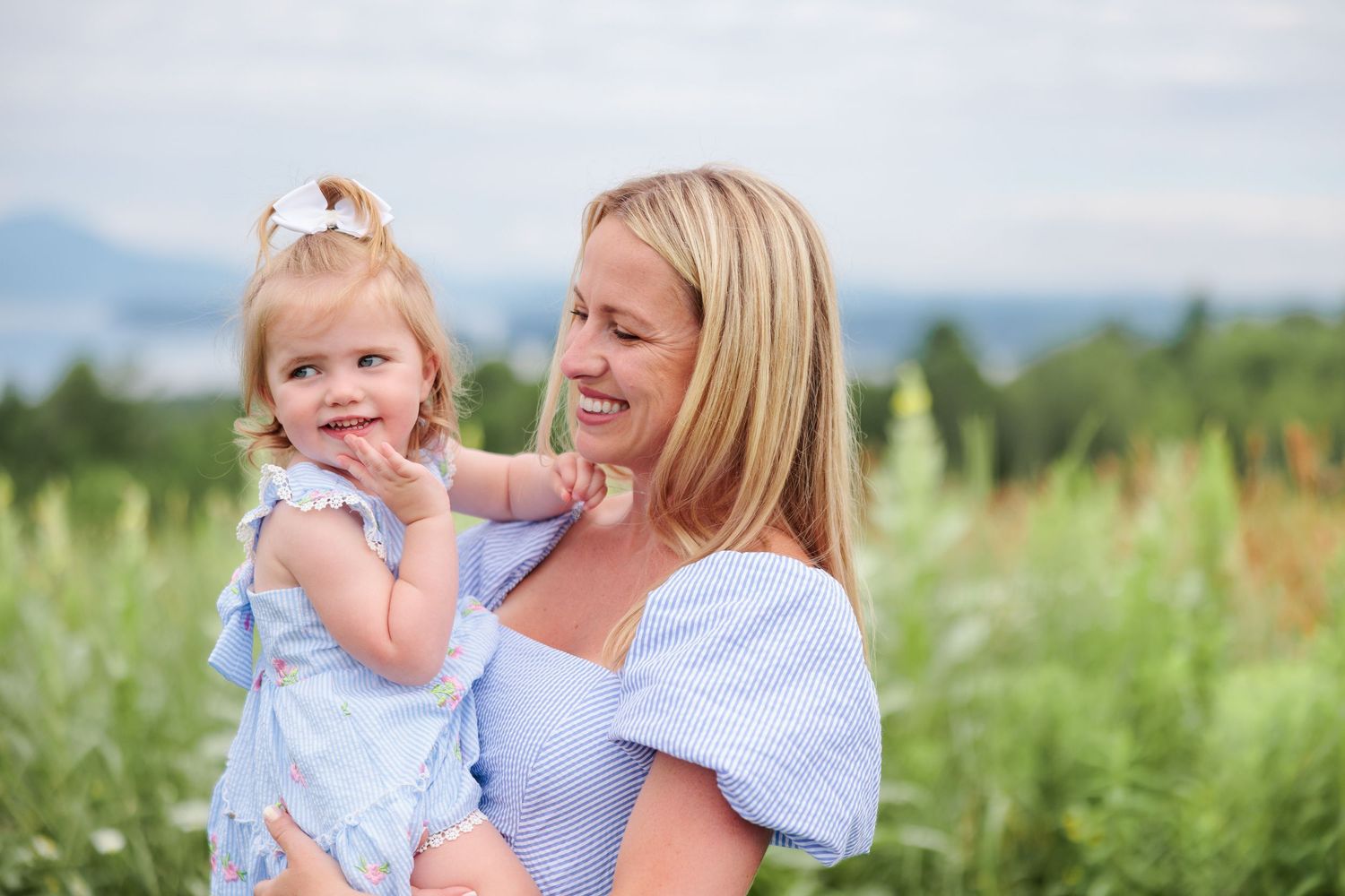 A blonde mother holds her toddler daughter in a green field, both wearing matching blue outfits.