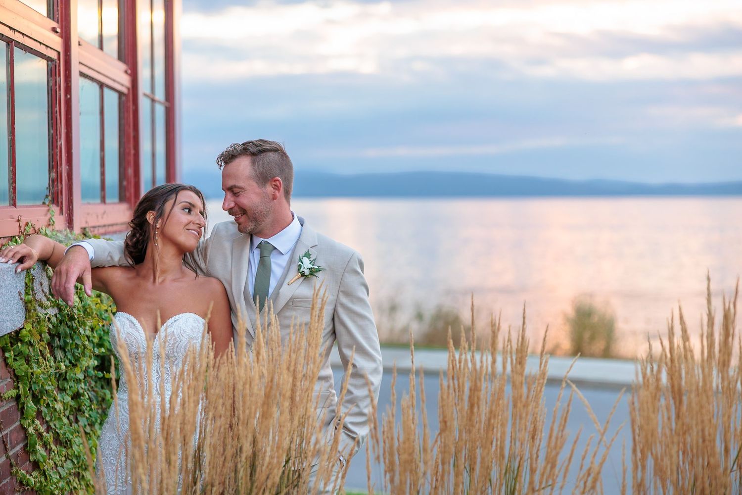 Bride and groom share a romantic sunset moment by the water, surrounded by tall golden grasses.
