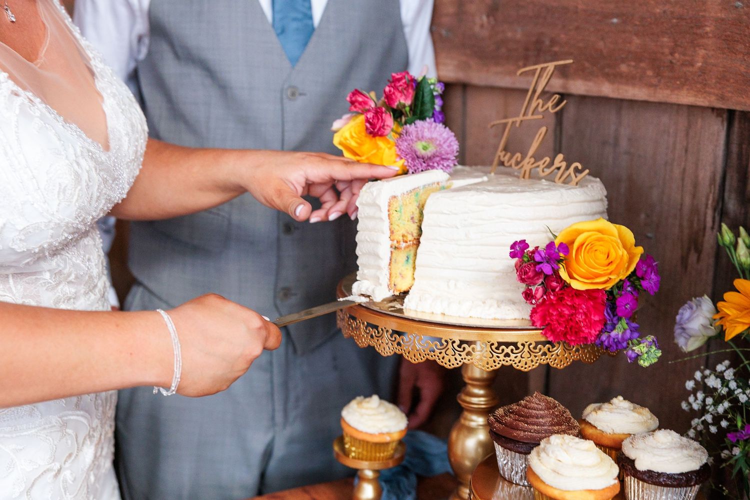 Bride and groom cutting white wedding cake adorned with colorful flowers on a gold stand.