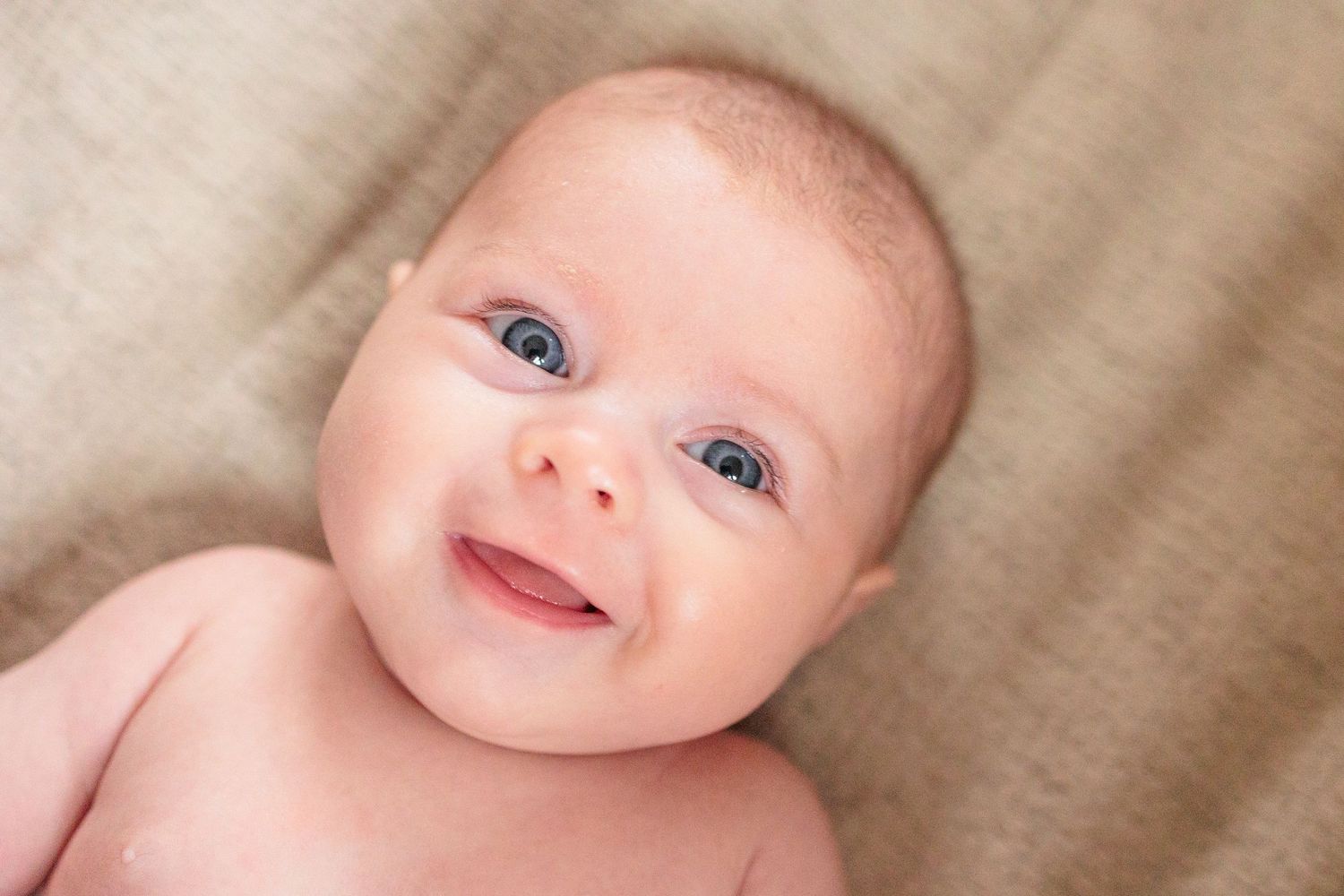 Adorable smiling blue-eyed newborn baby lying on soft beige blanket looking up at camera.