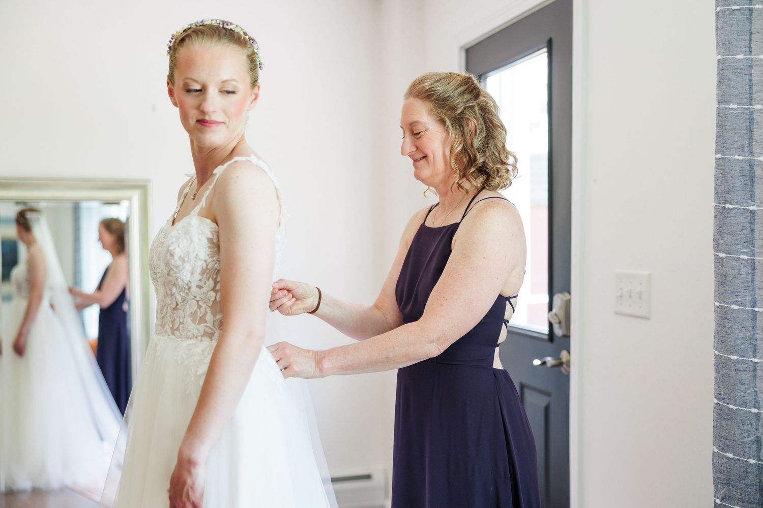 Bridesmaid in navy dress helping bride with lace wedding gown in bright dressing room.