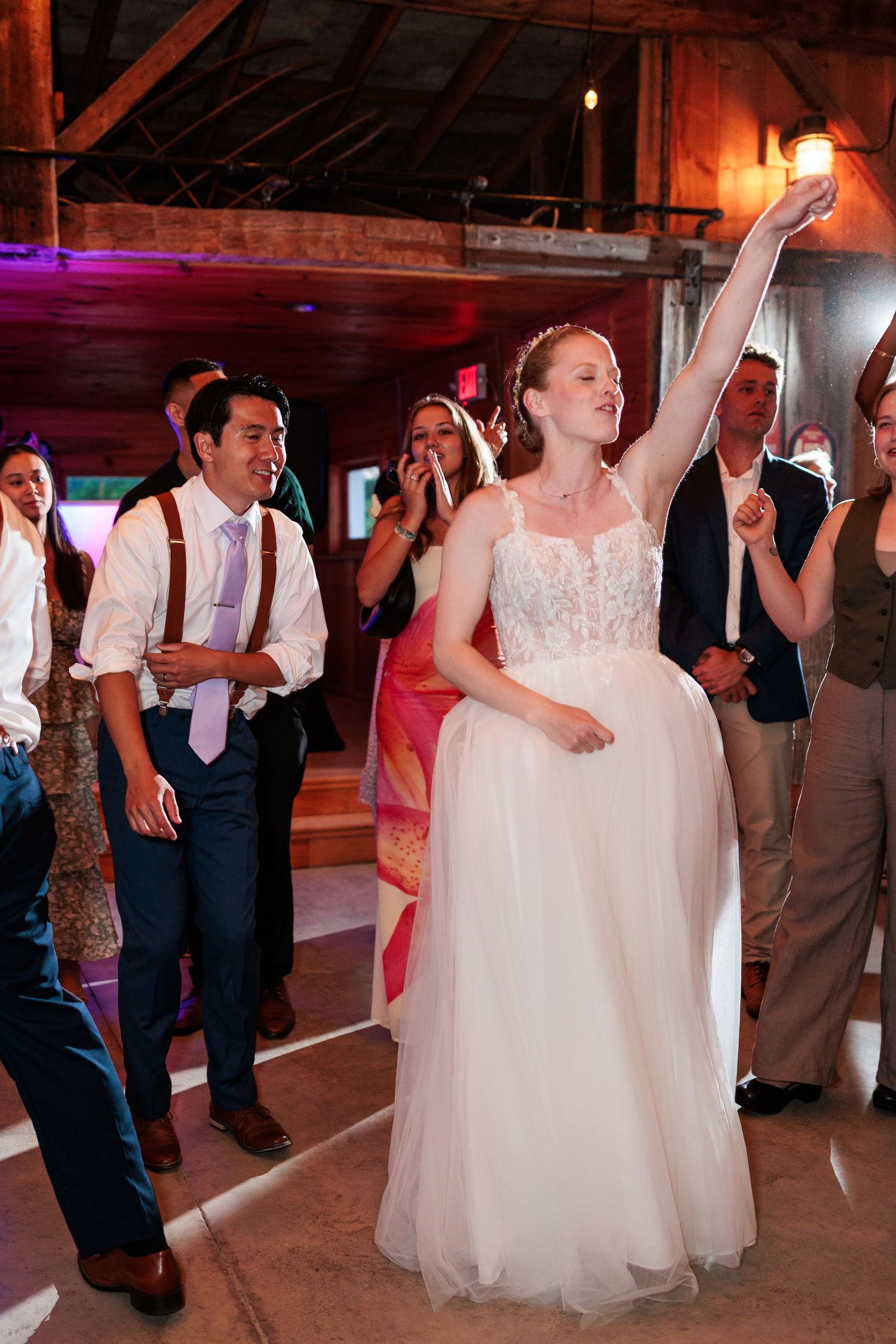 Radiant bride in white gown raises arm joyfully while dancing with guests at lively barn wedding reception.