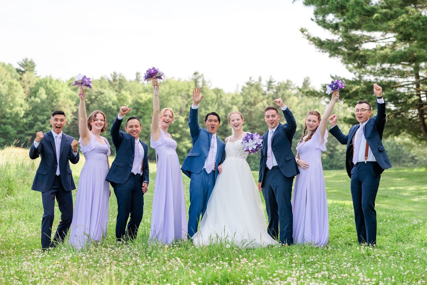 Joyful wedding party celebrates with raised arms in lush green outdoor setting.