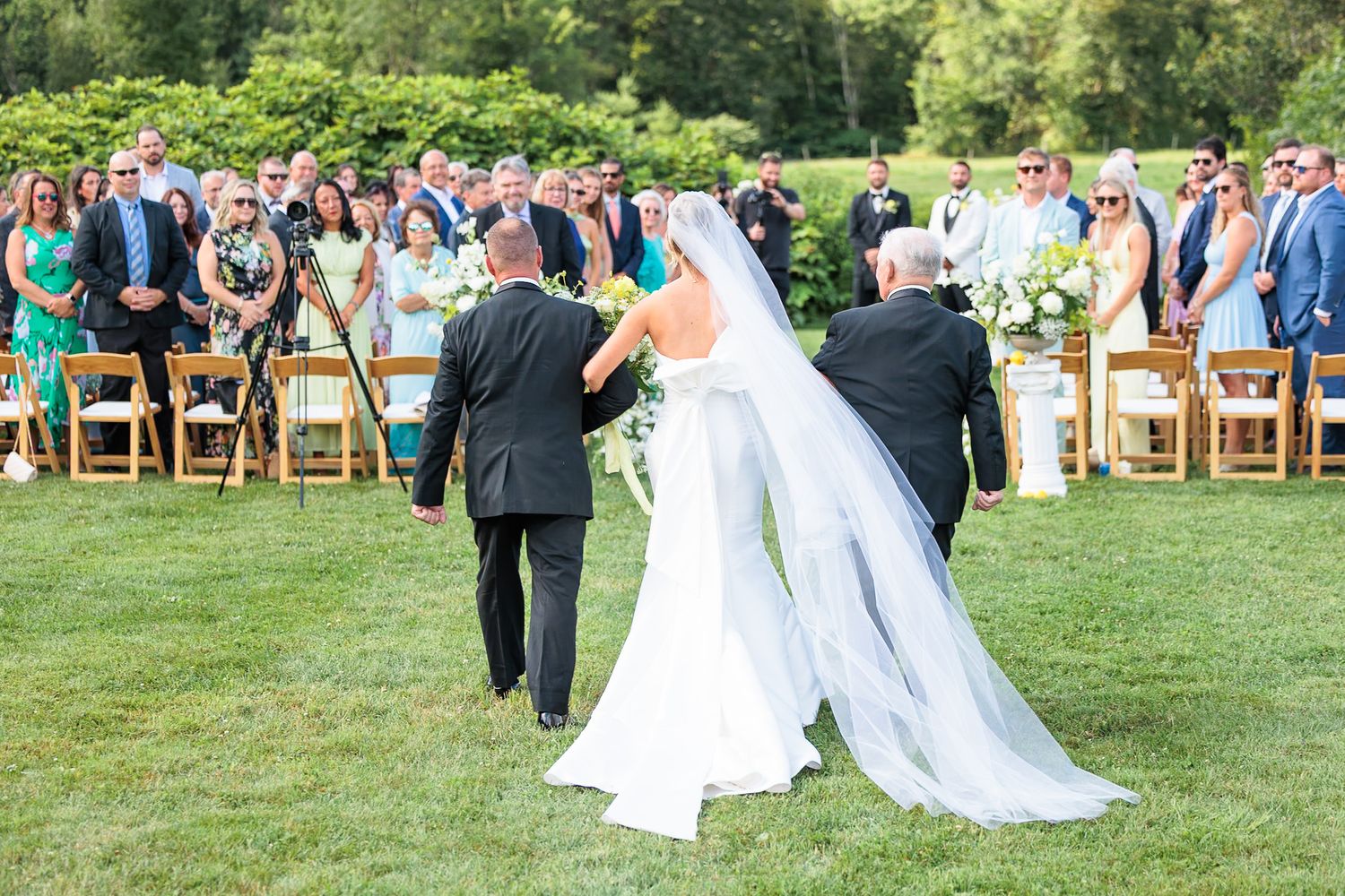 Bride in white gown and veil being walked down the aisle at outdoor ceremony.