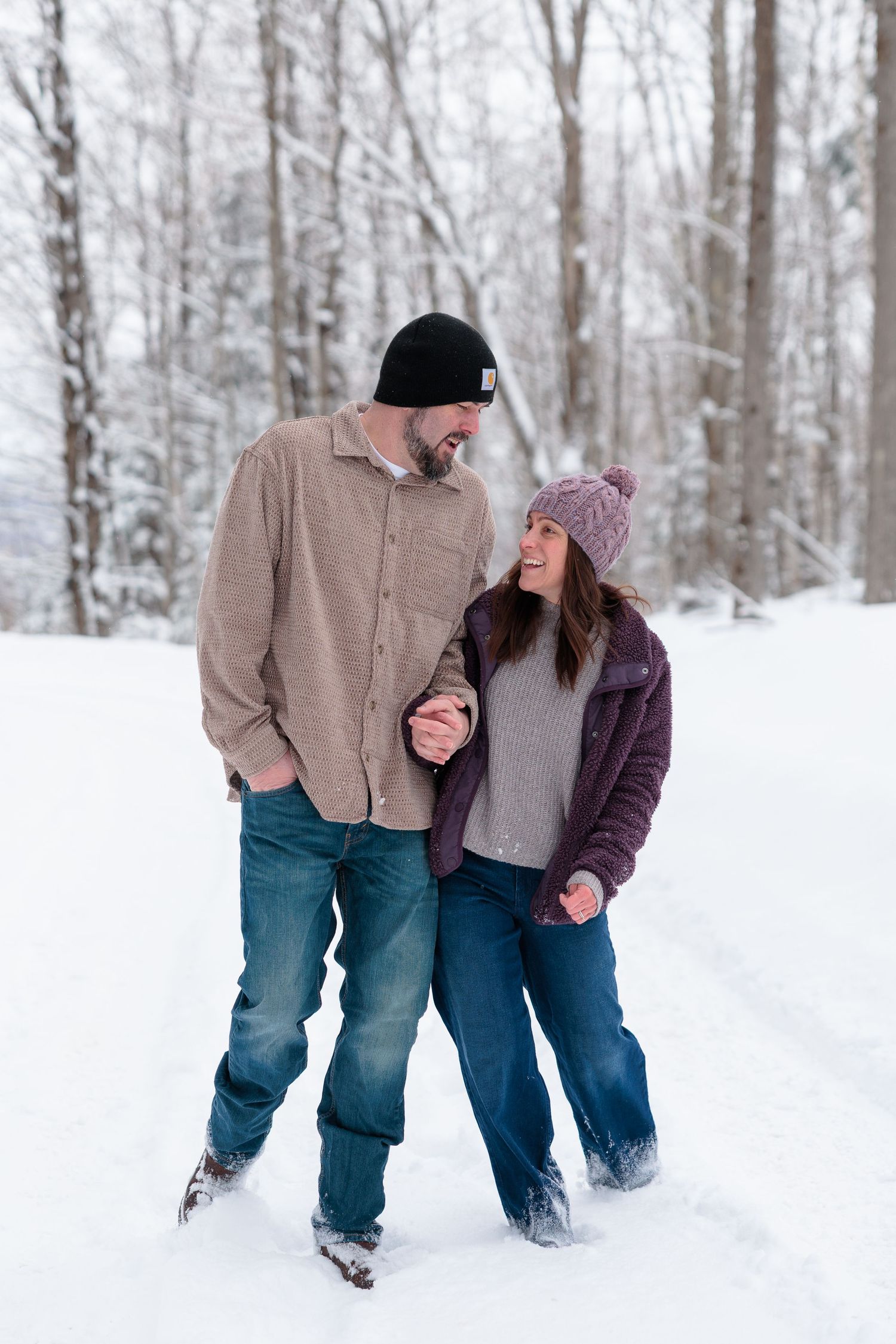 Couple holding hands and smiling at each other while walking through a snowy winter forest.