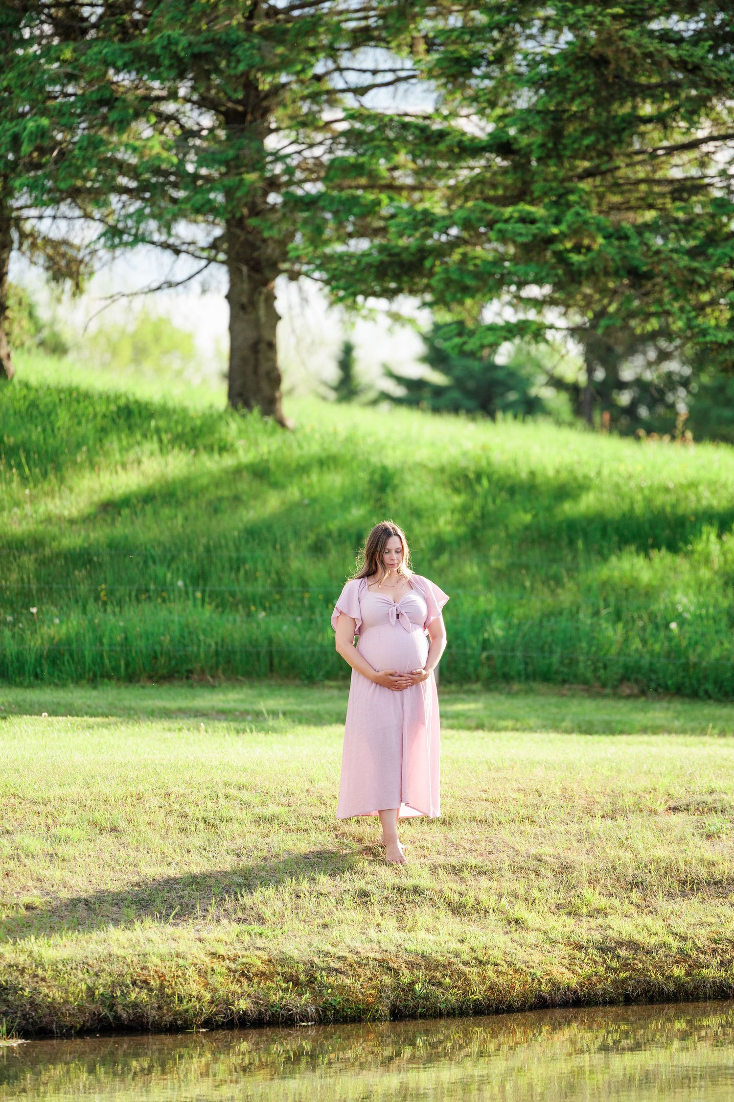 Pregnant woman in pink dress standing barefoot by a pond near tall trees.