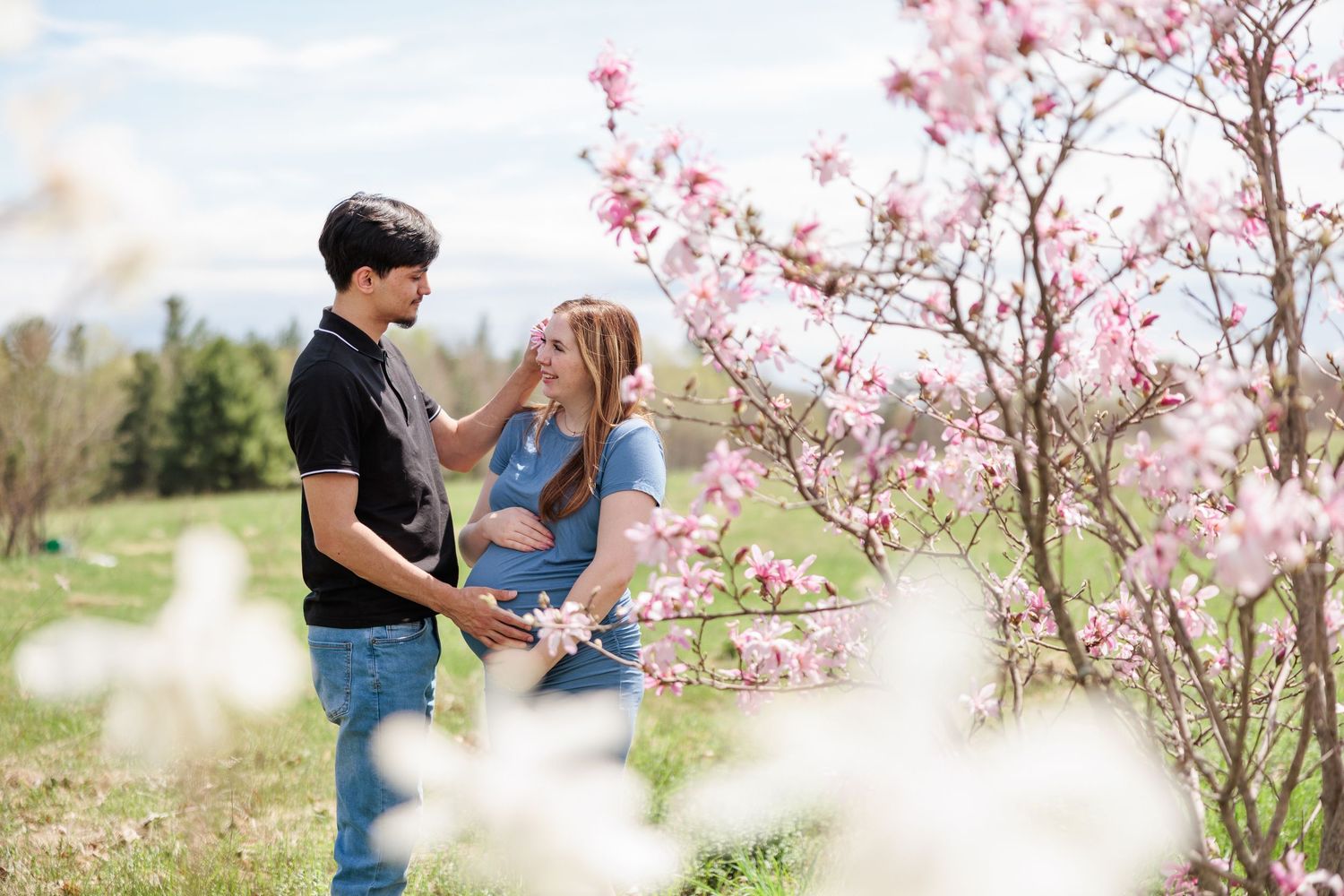 Couple holding hands near pink blossoming magnolia trees during a spring maternity photoshoot outdoors.