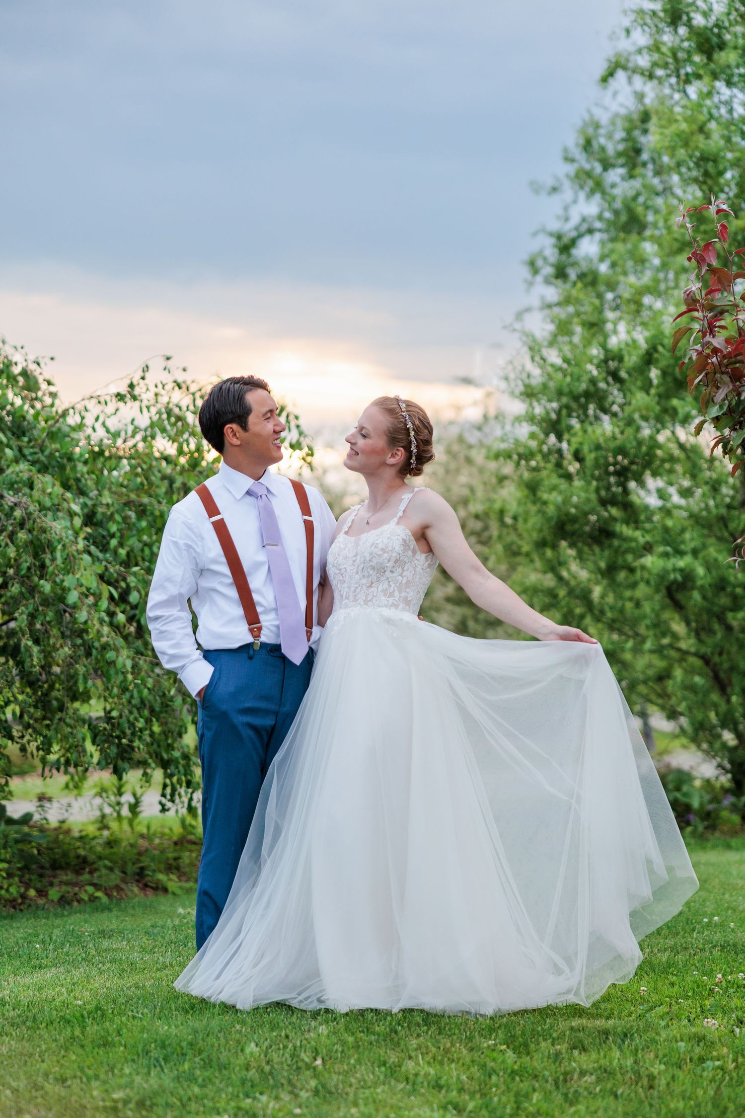 A bride in a white gown and groom in blue suit with suspenders share a romantic moment outdoors at sunset.