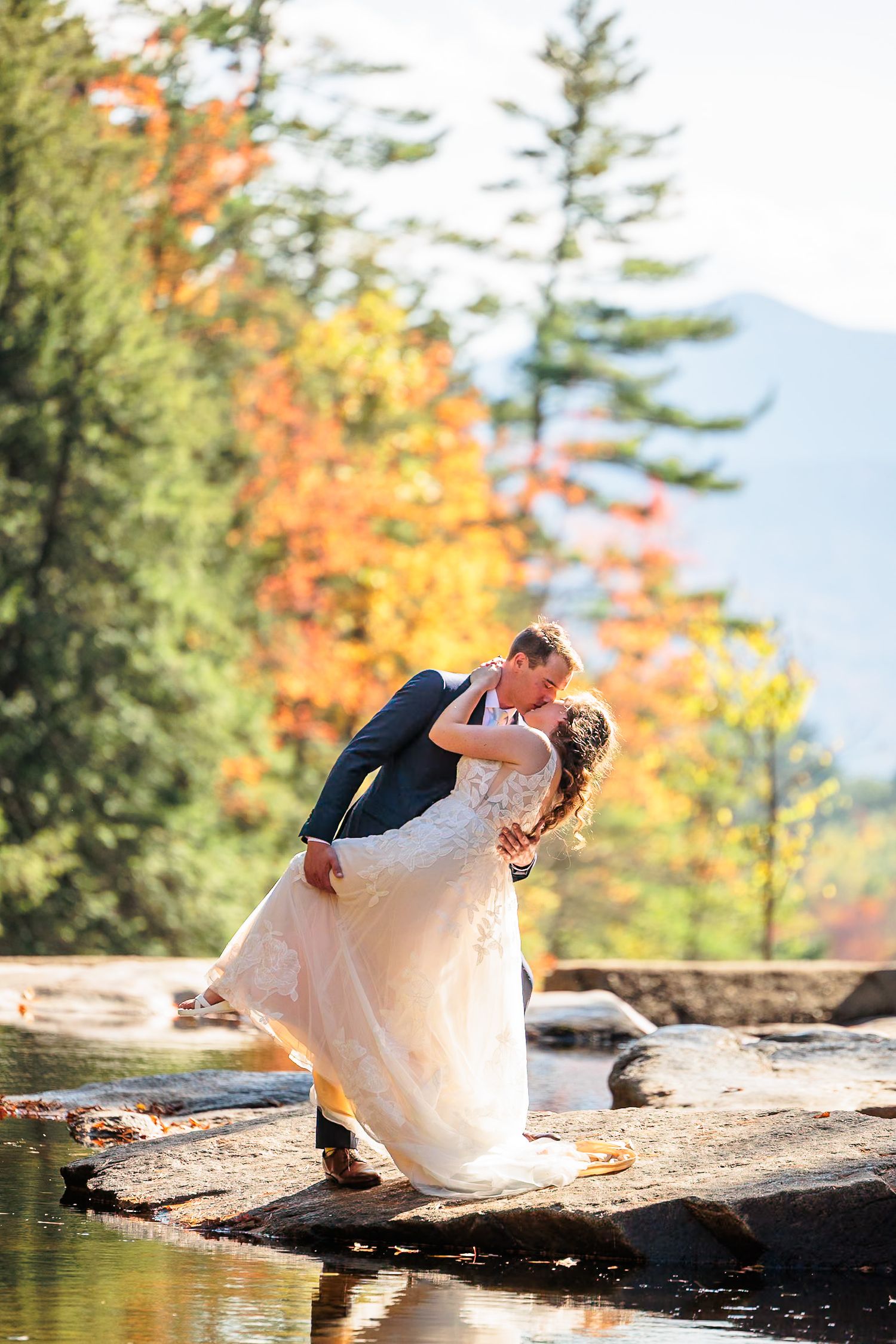 Bride and groom share a romantic dip kiss on rocks by a river surrounded by colorful autumn foliage.