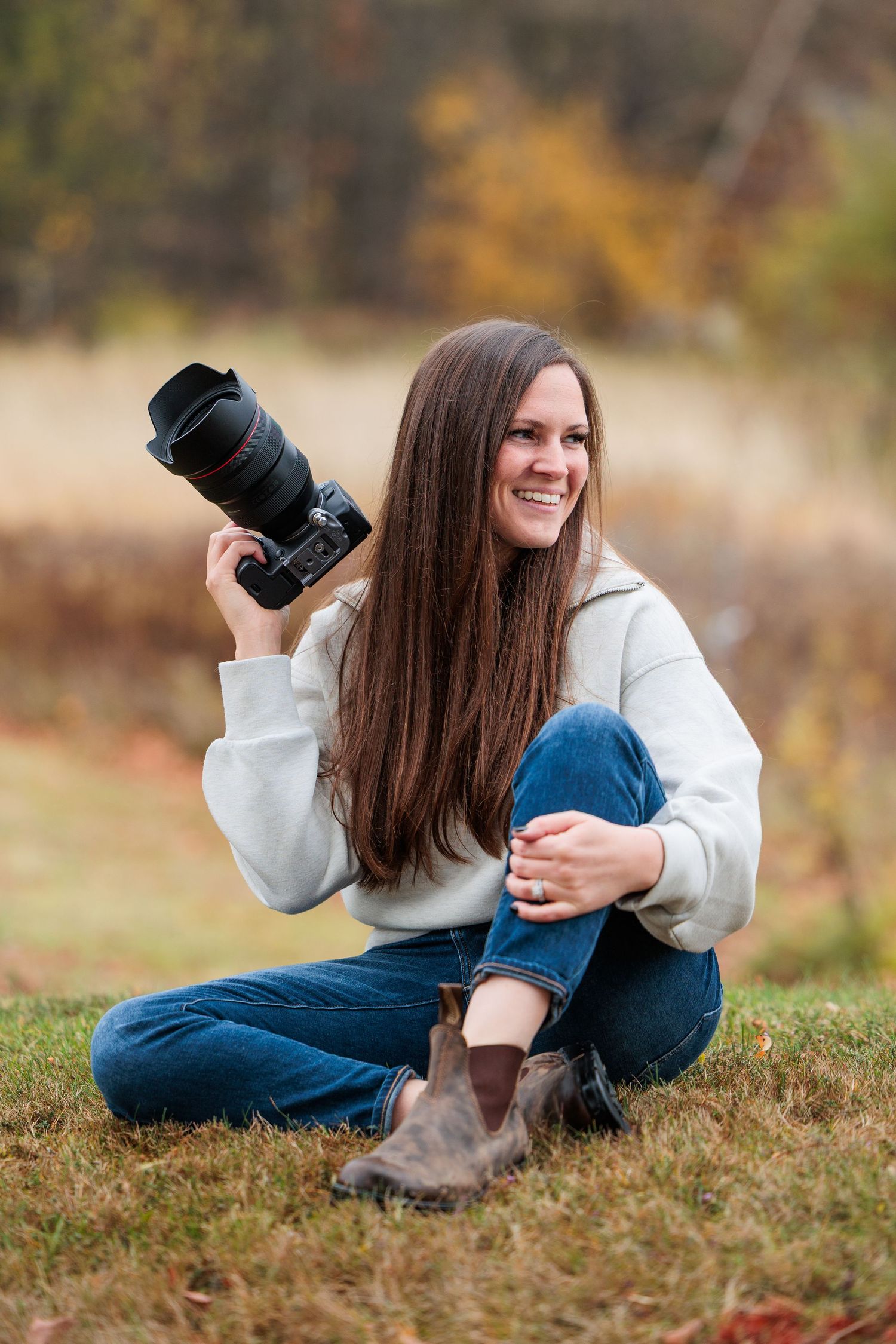 Smiling woman with long brown hair sitting on grass outdoors, holding a professional DSLR camera with large lens.