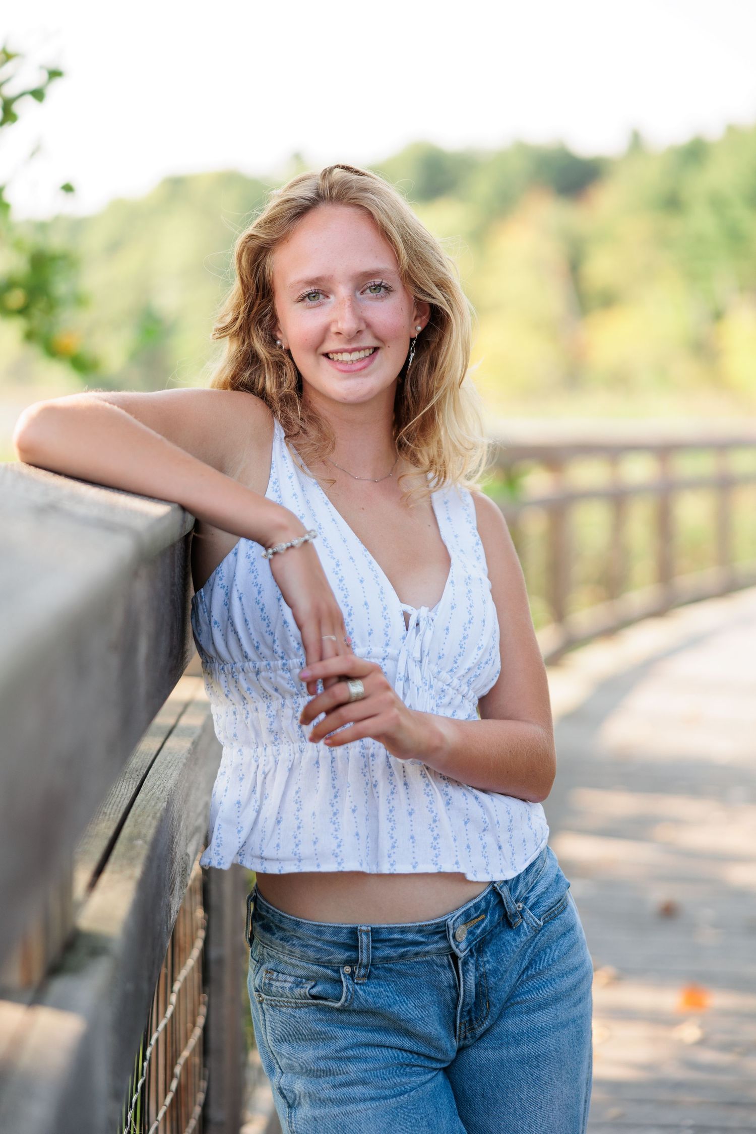 Smiling blonde teen girl in white crop top and jeans leaning on wooden bridge railing outdoors.
