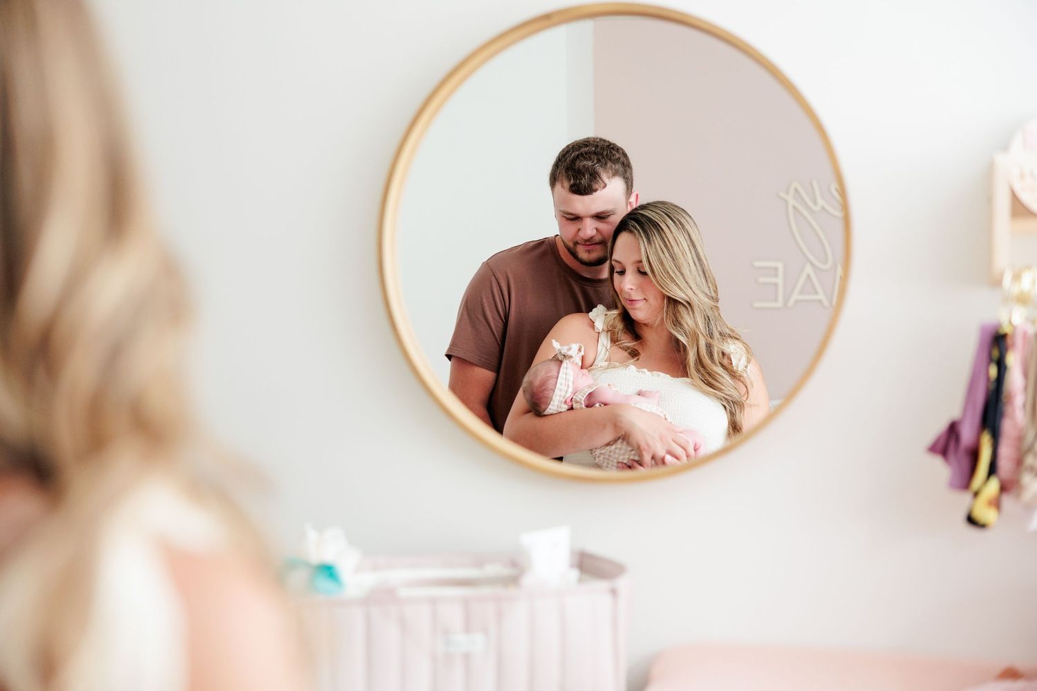 Couple with newborn baby reflected in round nursery mirror above white crib.