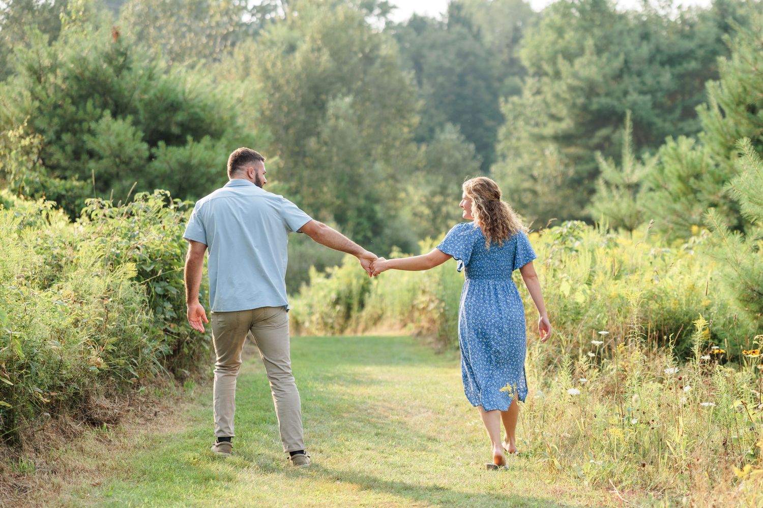 Couple walking hand in hand along a sunny green nature trail, viewed from behind.